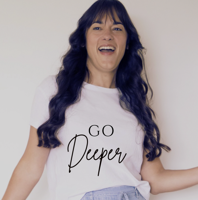 A woman with long, wavy, dark blue hair, wearing a white t-shirt with the text 'Go Deeper', smiling and standing against a plain background.