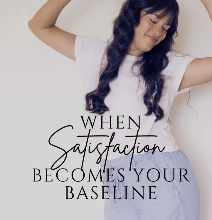 A woman with long dark wavy hair smiling with her eyes closed, wearing a white t-shirt and light-colored pants, with the text 'When Satisfaction Becomes Your Baseline' overlaid on the image.