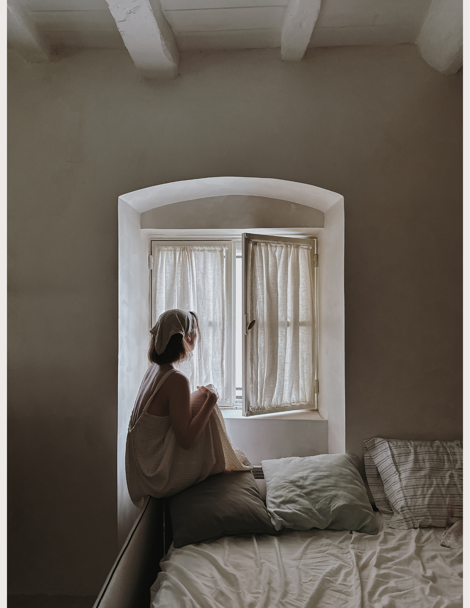 A woman sitting on the corner of a bed near an arched window, gazing outside. The bed has pillows and is in a softly lit room with beige curtains.