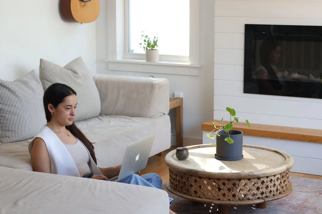 A woman sitting on the floor next to a beige couch, working on a silver laptop. There is a round wooden coffee table with a potted plant and a small dark decorative item in front of her. A window behind her lets in natural light, and a fireplace with a white wooden mantel is visible on the wall to her right.