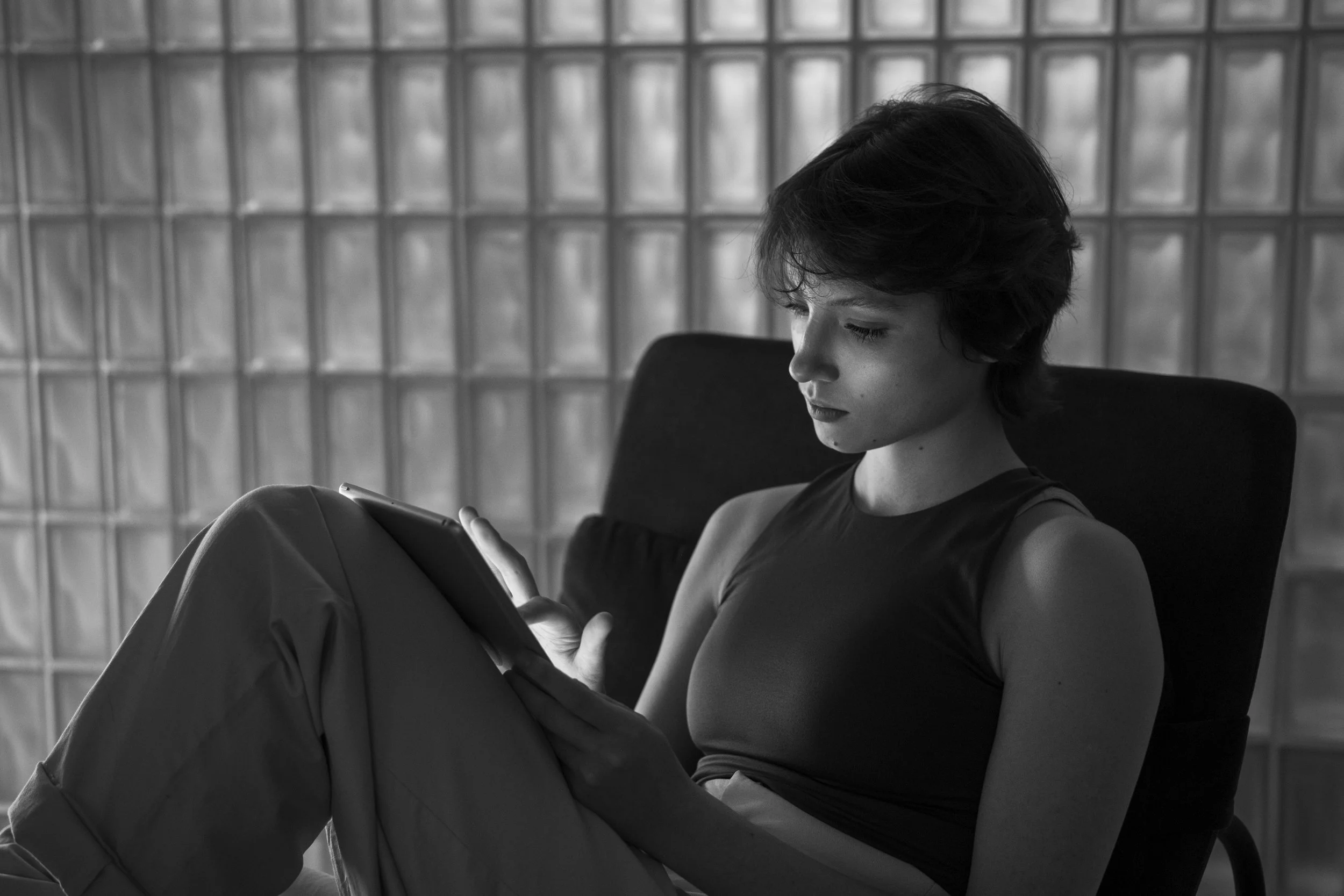 A woman with short dark hair sitting on a chair, looking at a tablet or phone, in front of a background with glass blocks.
