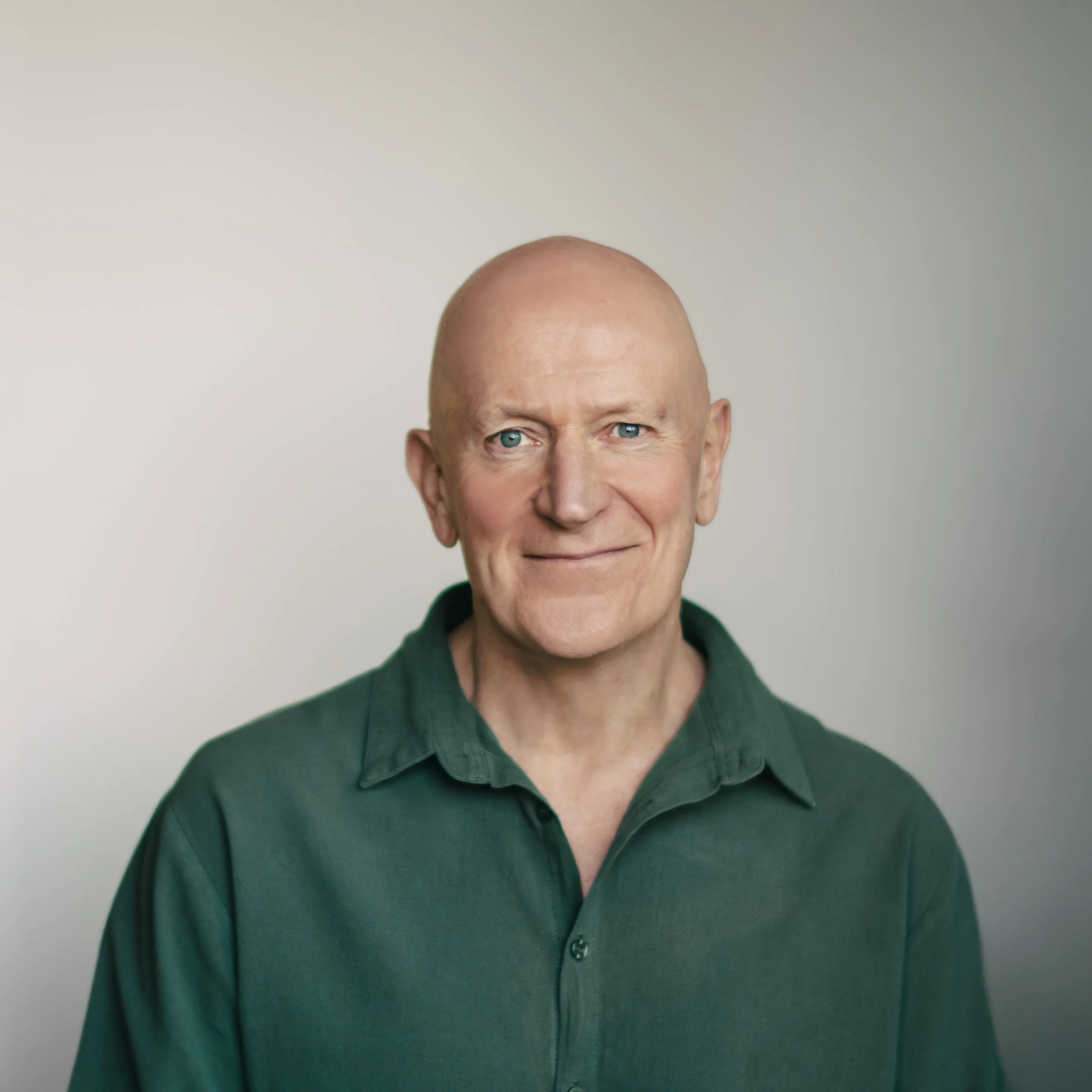 A middle-aged man with a bald head, blue eyes, and light skin, wearing a white dress shirt, smiling softly while sitting against a plain, light-colored background.
