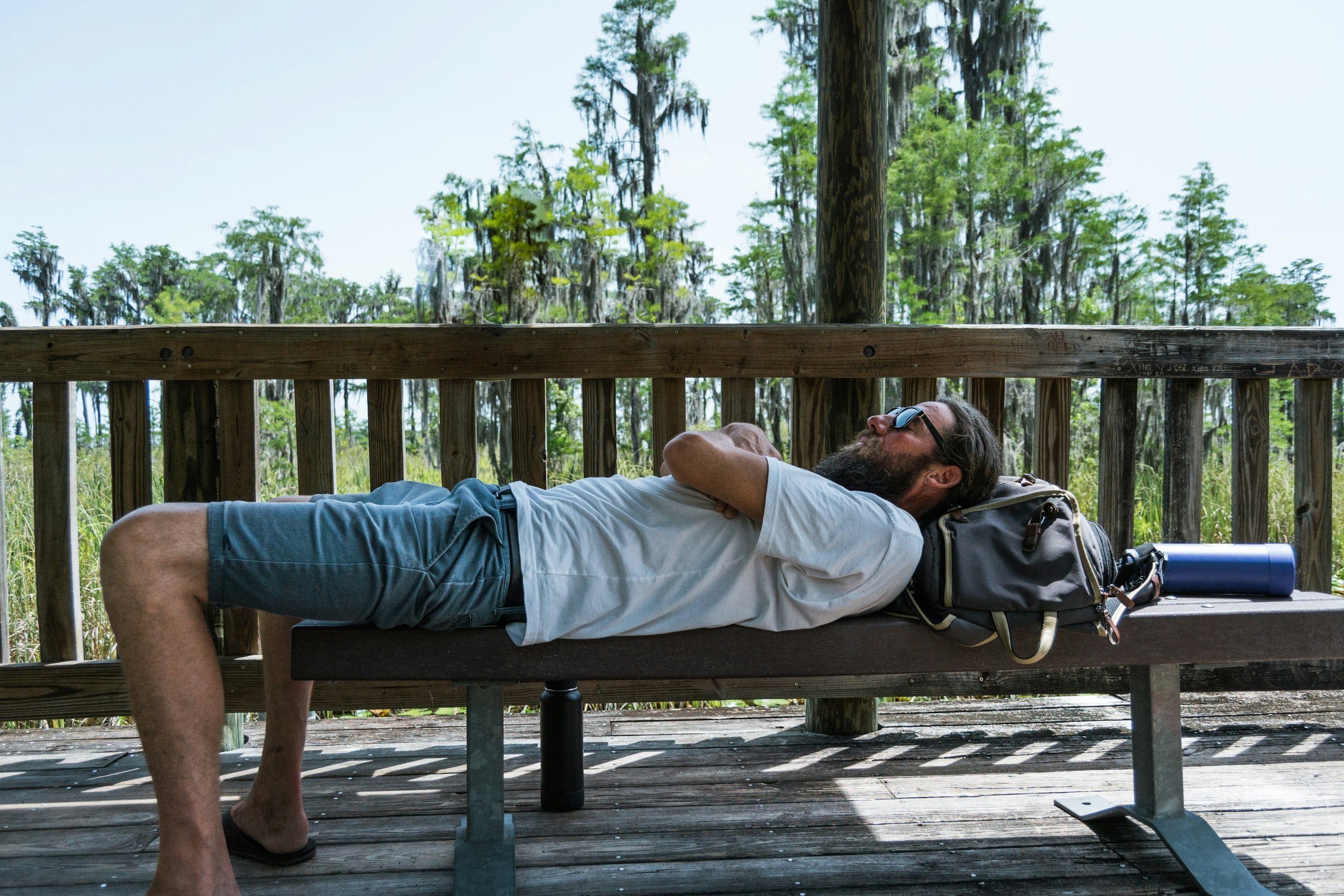 A man with sunglasses, a beard, and a white t-shirt is lying on his back on a bench on a wooden deck, with his arms crossed over his chest, in a forested area with tall trees and greenery.