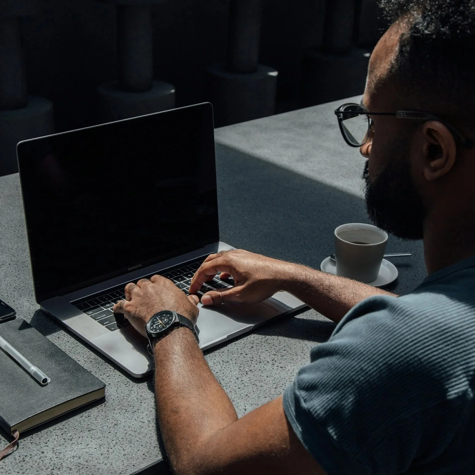 A man working on a MacBook at an outdoor table with a coffee cup, notebook, and pen, wearing glasses and a watch.