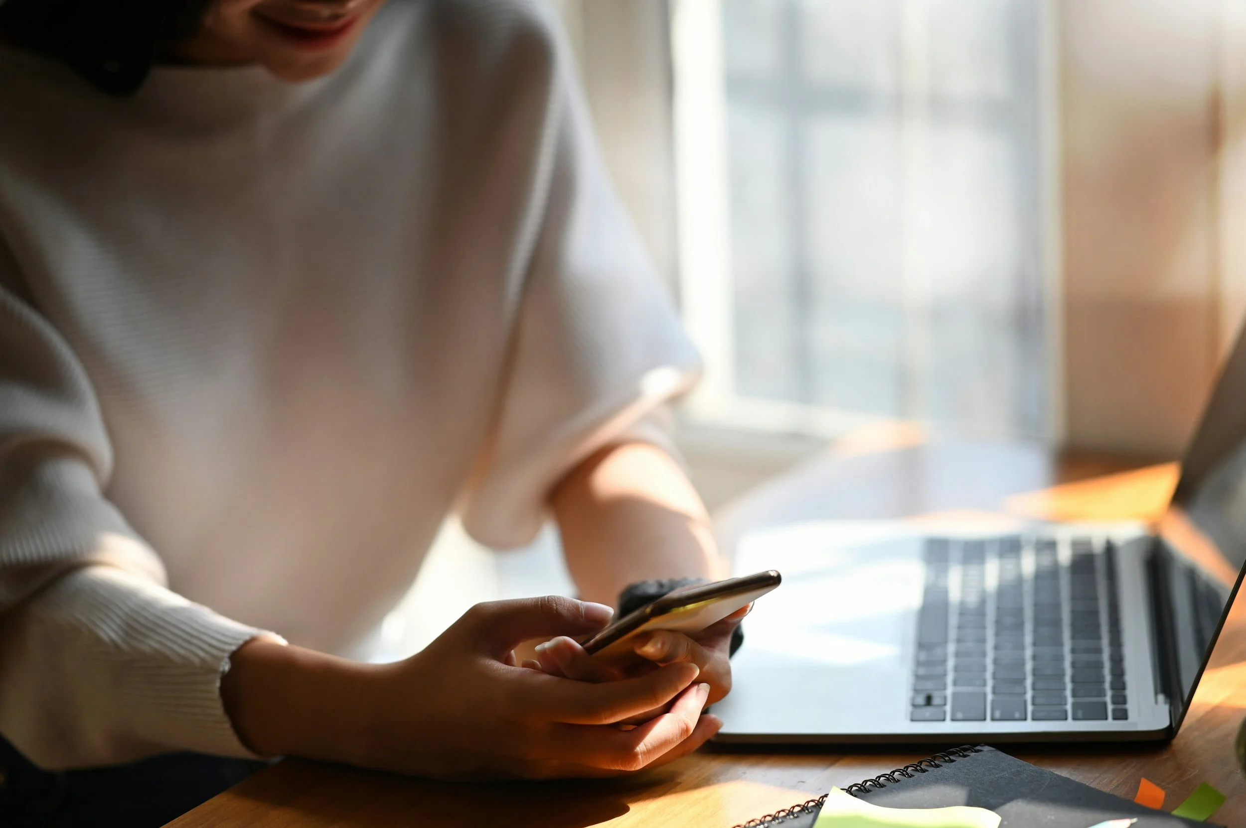 A person holding a smartphone while sitting at a desk with a laptop and papers, near a window letting in natural light.