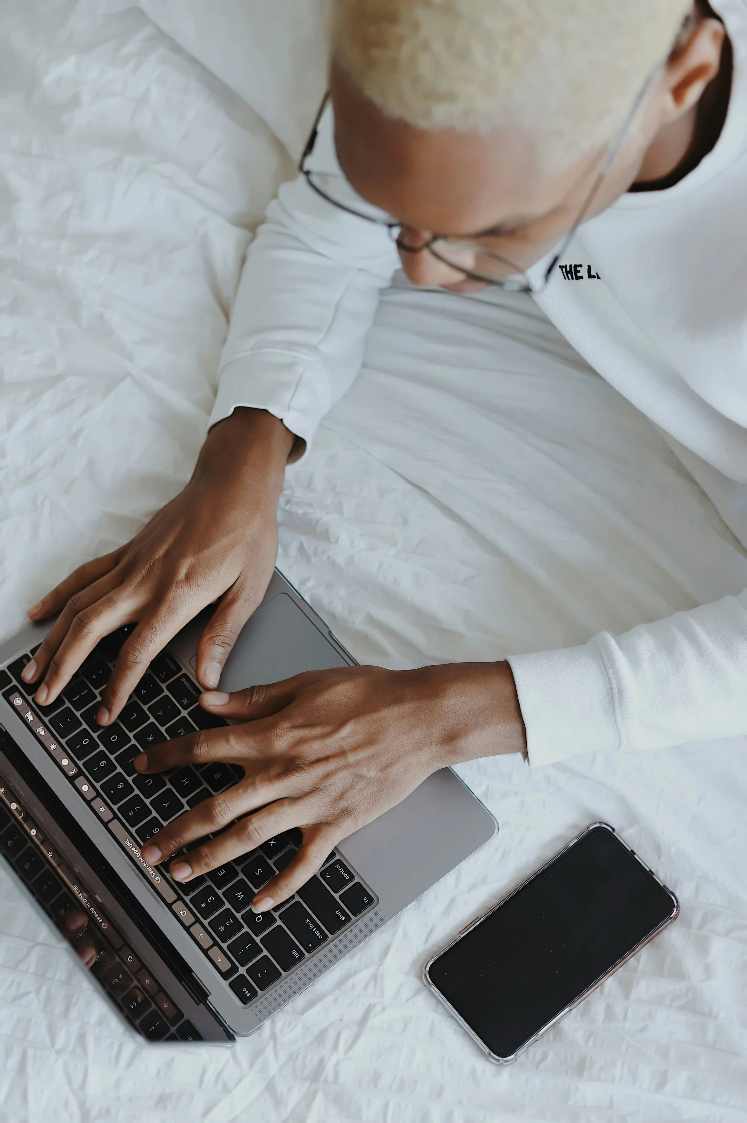 A person with short blonde hair and glasses wearing a white shirt is typing on a laptop, with a smartphone beside it, on a white bed.