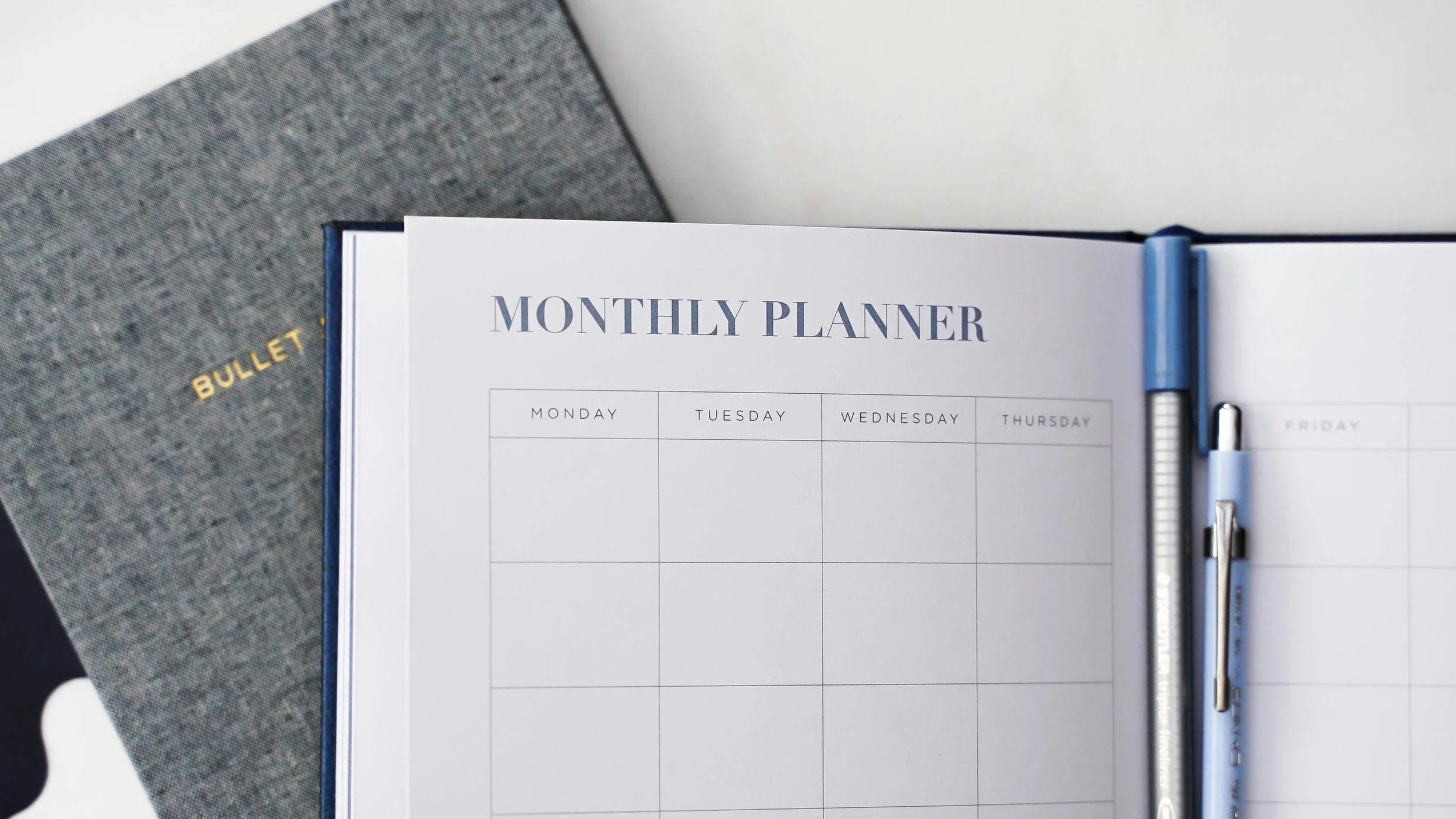Open monthly planner with blank weekly pages, titled 'Monthly Planner,' with two pens clipped to the right side.