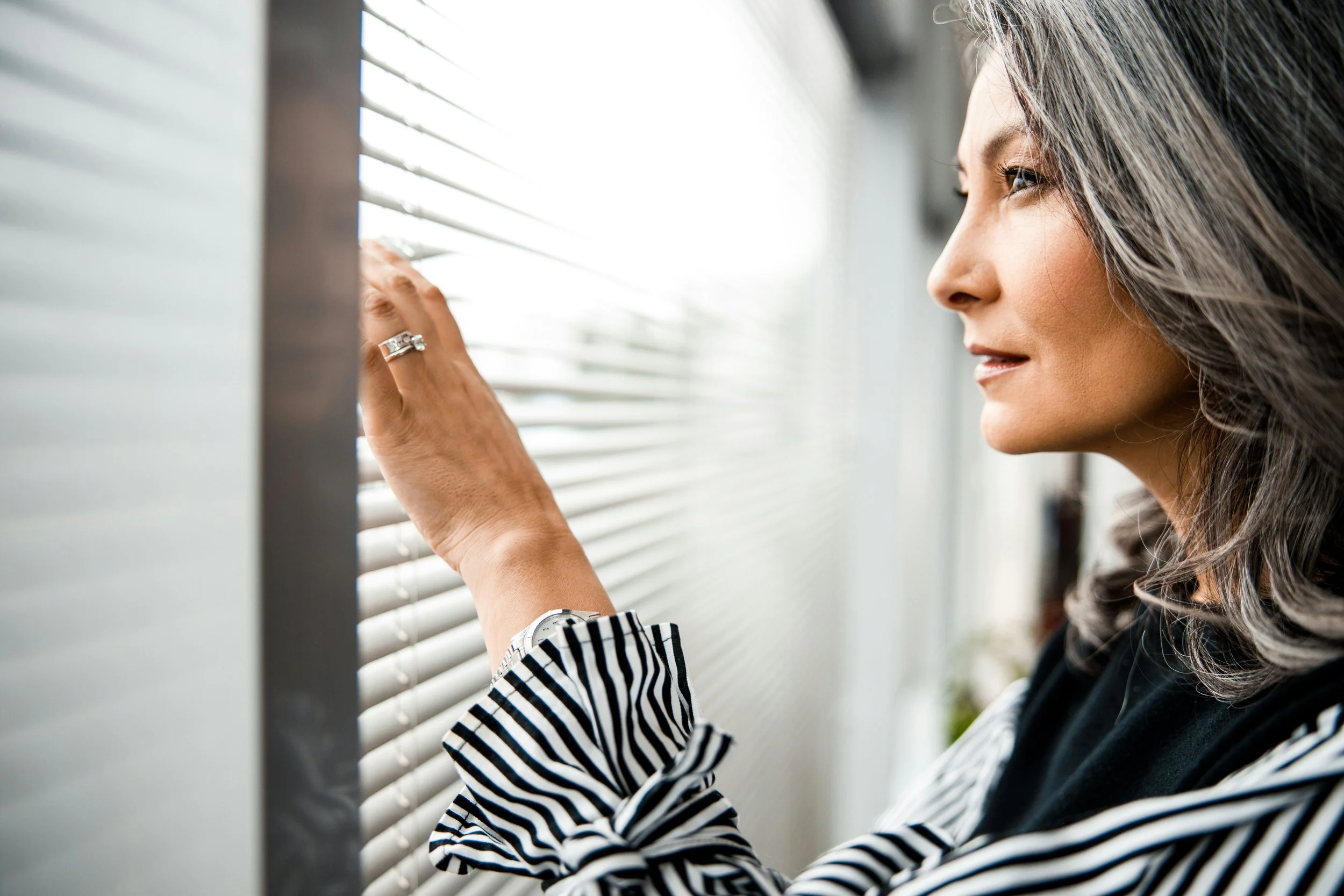 A woman looking out a window with horizontal blinds, her hand touching the blinds, wearing a striped sleeve, watch, and ring.