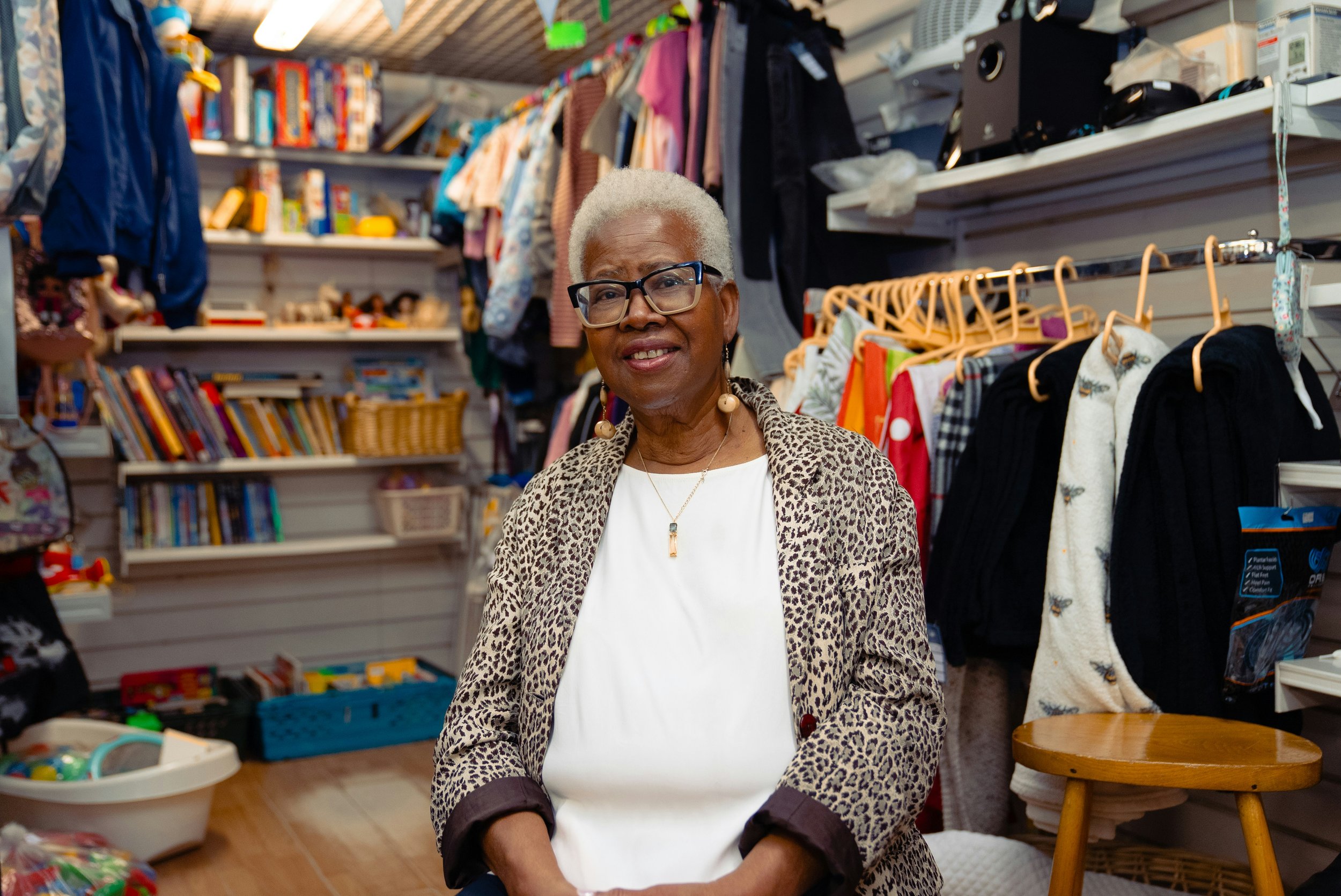 An elderly woman with short gray hair, glasses, and wearing a leopard print blazer and white top, sitting in a thrift store surrounded by clothing, books, and toys.