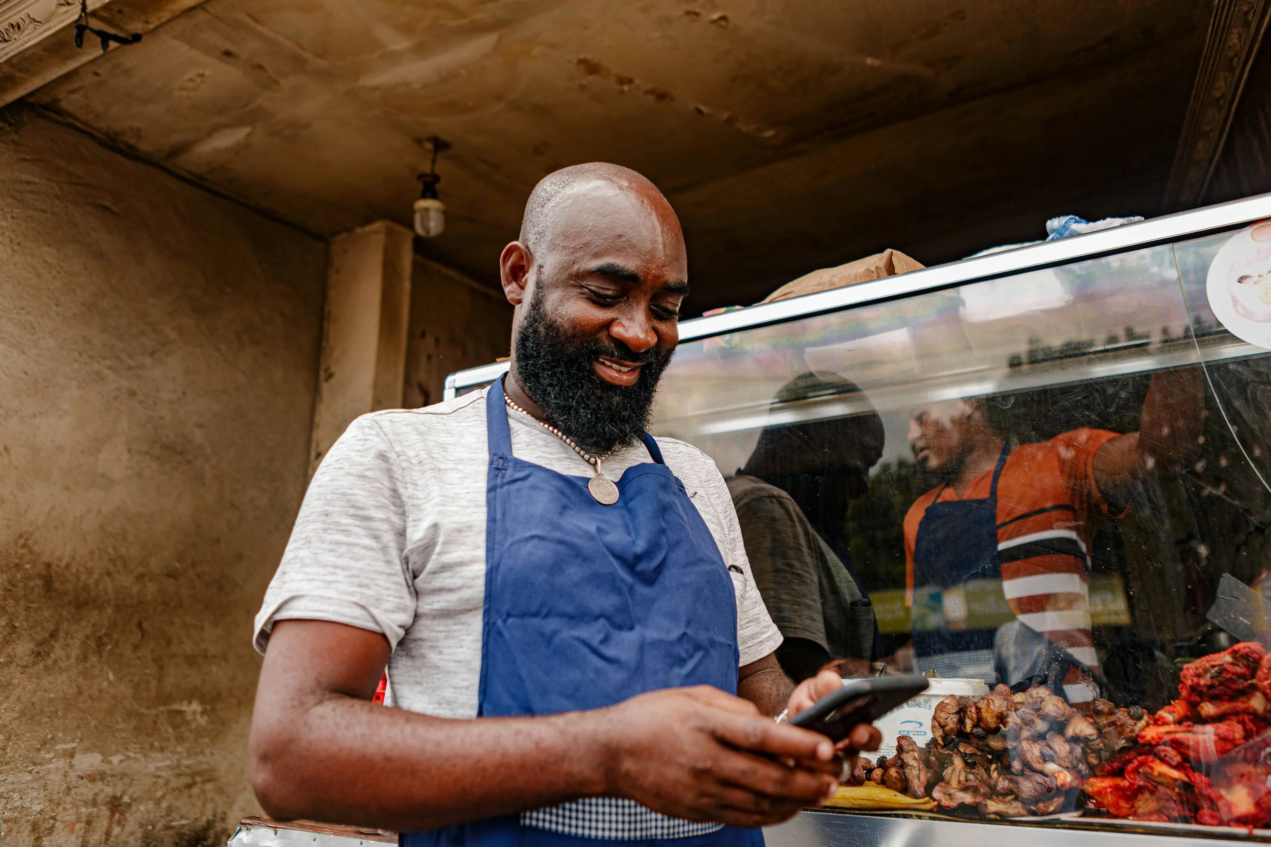 Man with a beard wearing a blue apron and necklace, smiling and looking at his phone inside a food stall with grilled meat.