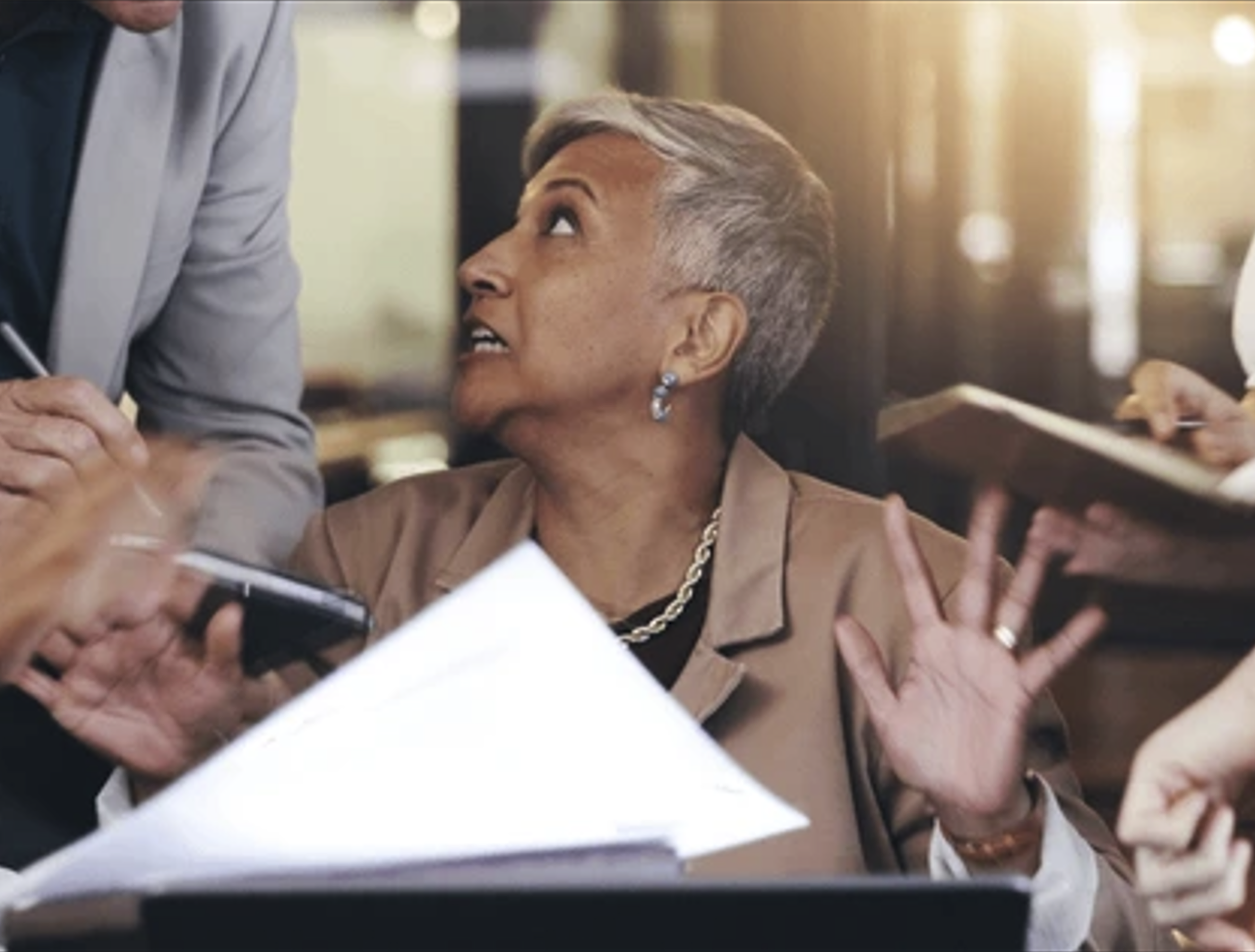 An older woman with short gray hair, wearing earrings, a necklace, and a brown blazer, appears to be surprised or upset while seated at a desk, with people around her in a professional setting.