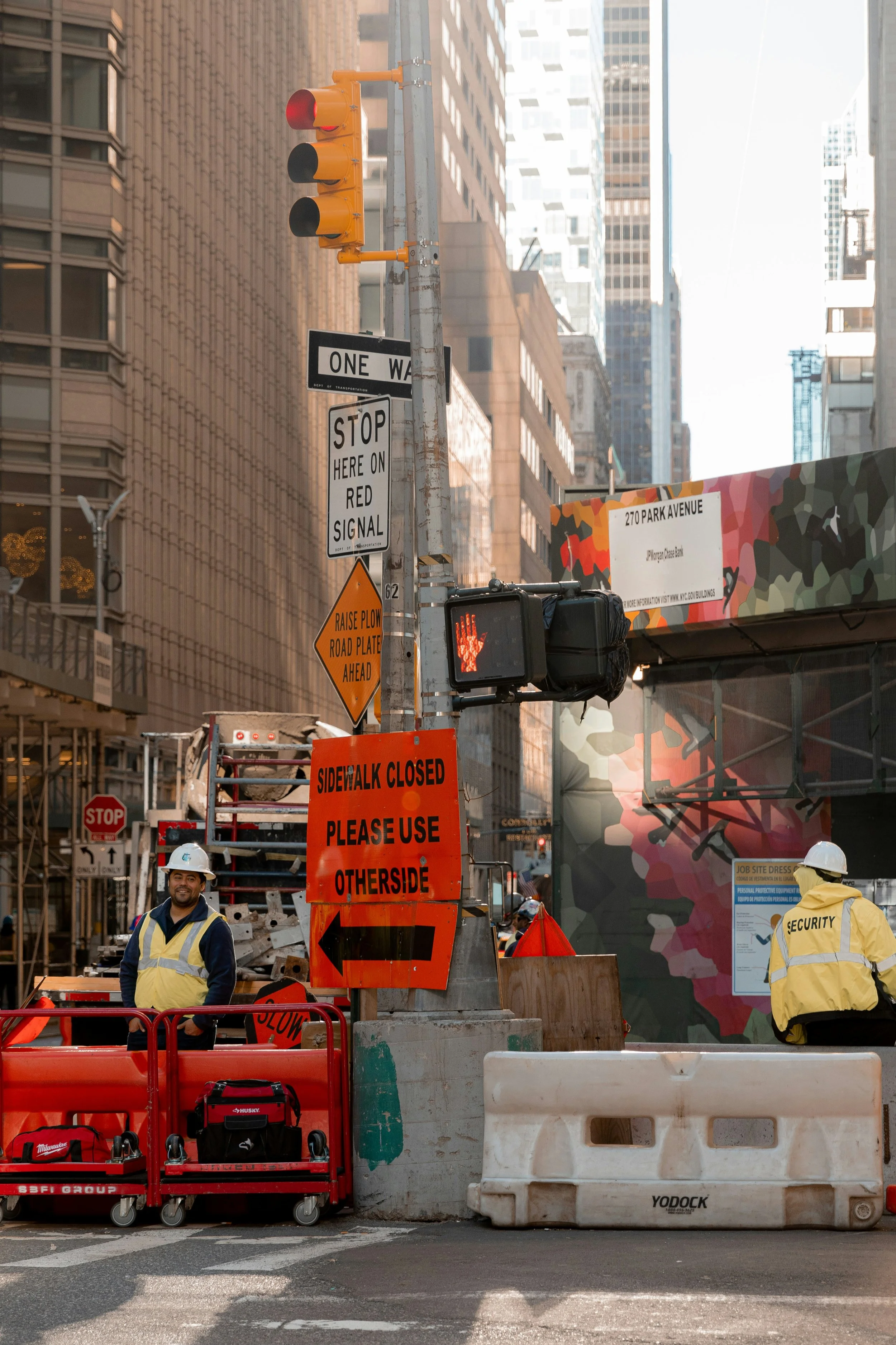 Construction workers in yellow safety vests and helmets working on a city street with traffic signs, barriers, and a traffic light.