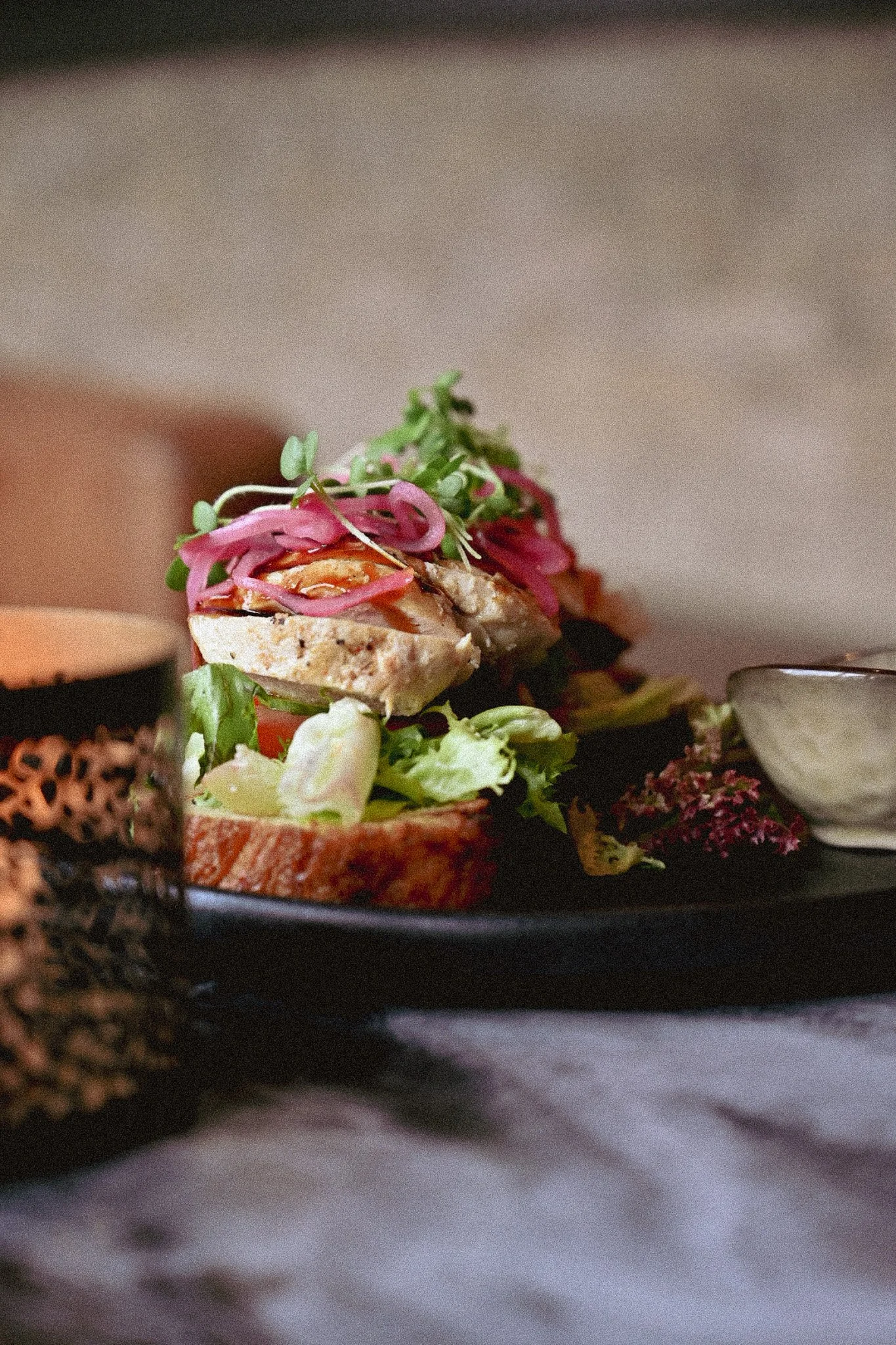 Nærbilde av en tallerken på et marmorbord, med kyllingsmørbrød, grønnsaker, og en liten tallerken med saus, serveres på en svart tallerken.