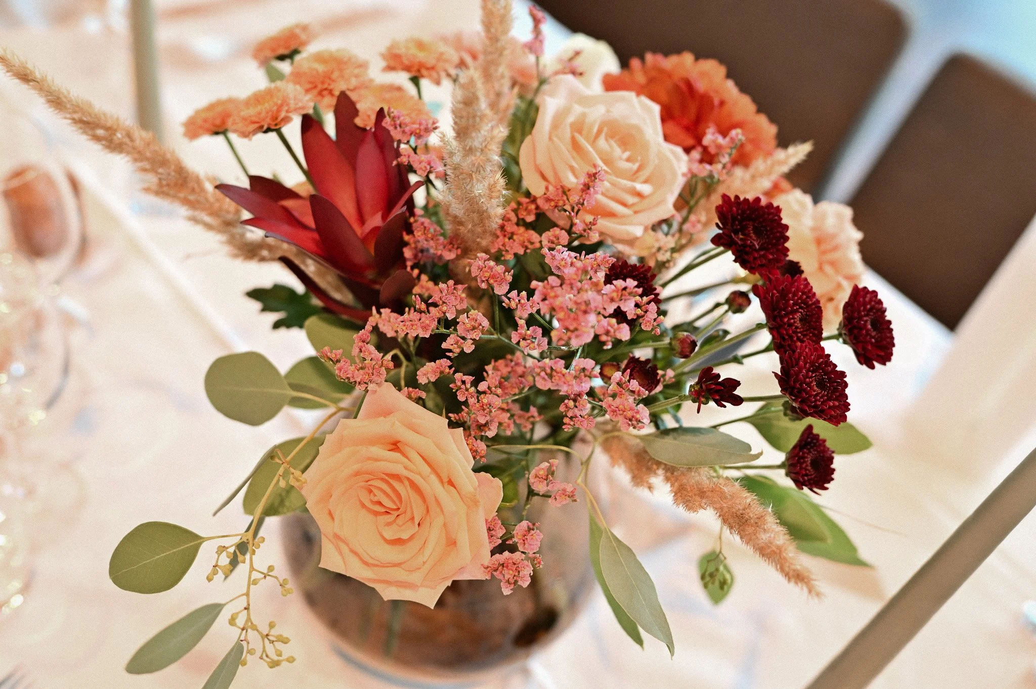 En blomsterbukett med rosa roser, røde og rosa småblomster, grønne blader og lys rosa Pampasgras i en glassvase.