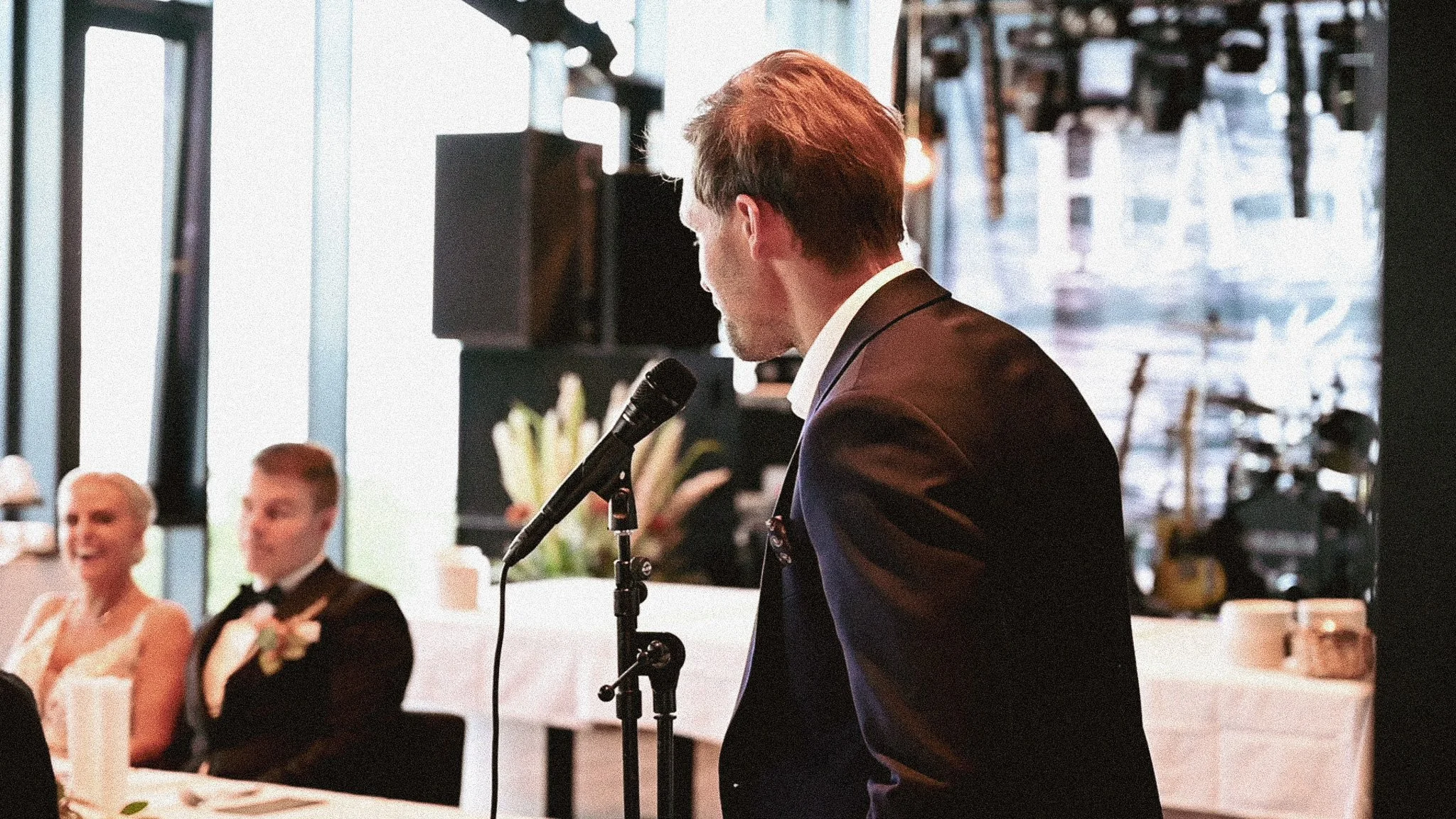 En mann i dress som snakker i mikrofon ved et bryllup. I bakgrunnen er en brud og en brudgom som smiler.