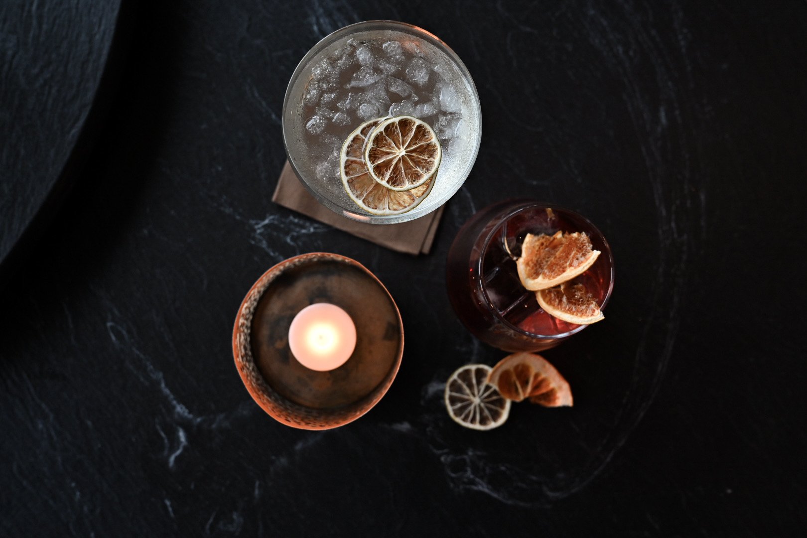 Høyt glass med en tørket sitron skive og isbiter, et lite lys i en kopp og et glass med en drink med sitronbiter, alle plassert på en mørk overflate.