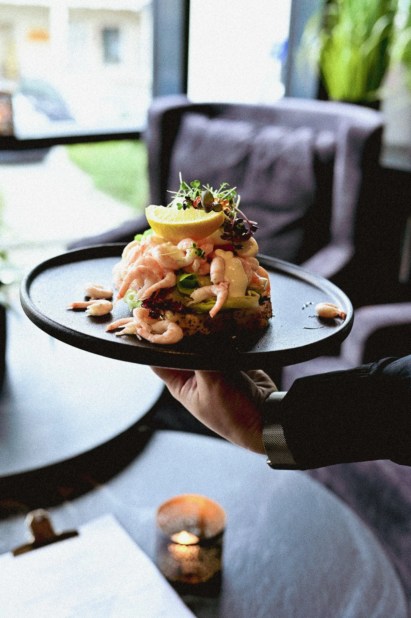 Person som holder et svart serveringsfat med smørbrød med reker, sitronskiver og garnityr.