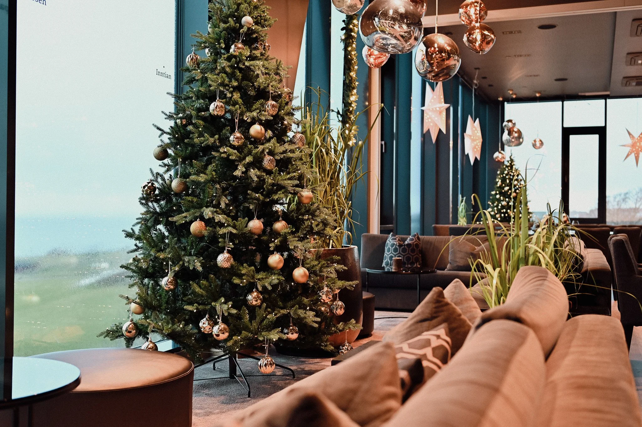 Innendørs juledekorasjon med pyntet juletre, julelys, stjerner og ornamenter i et rom med store vinduer og moderne møbler.