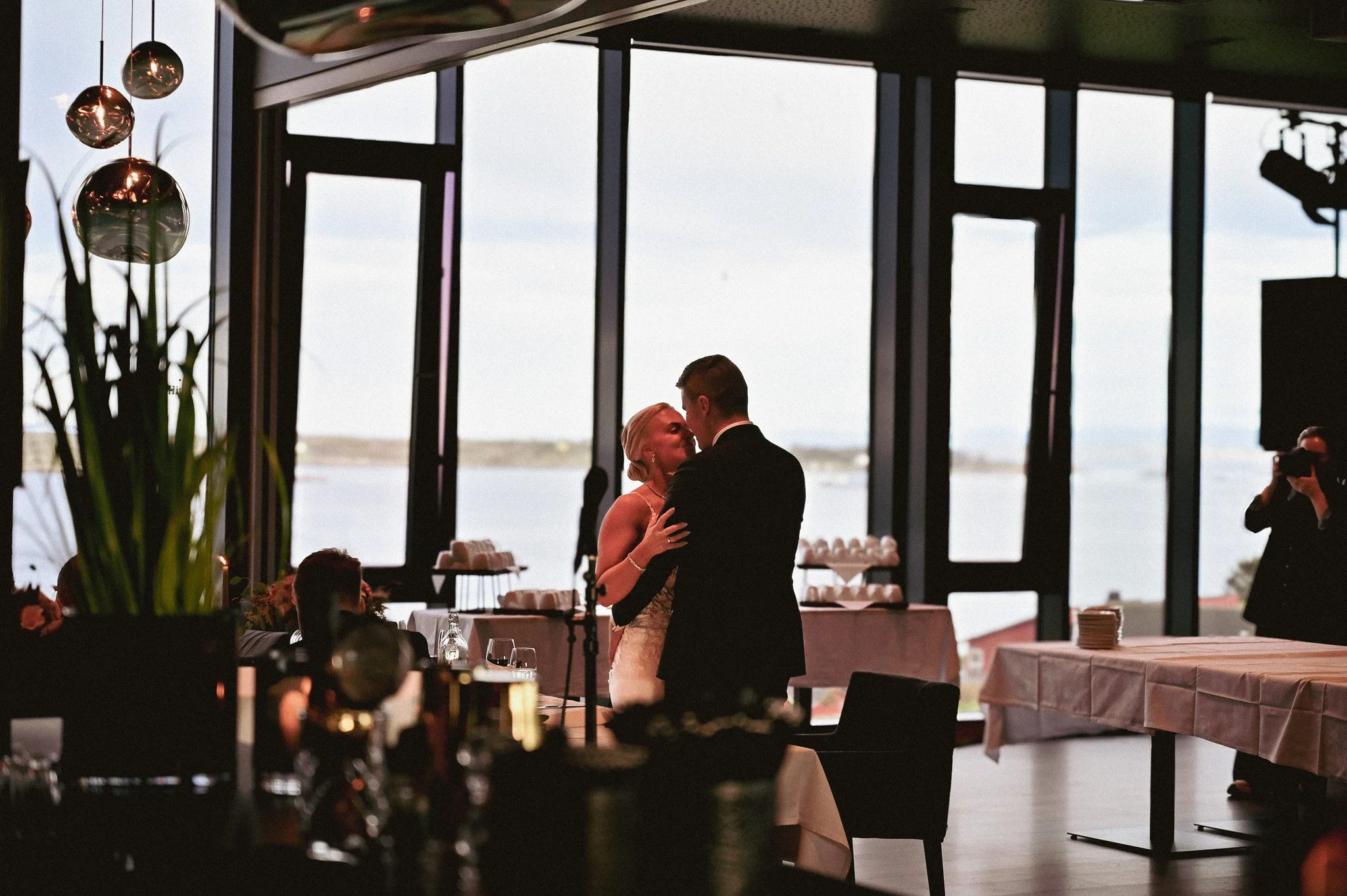 Et brudepar danser i et stort rom med store vinduer som viser utsikt over vannet. Det er dekorasjon og bordpynt på bordene, og en fotograf tar bilde av dem.