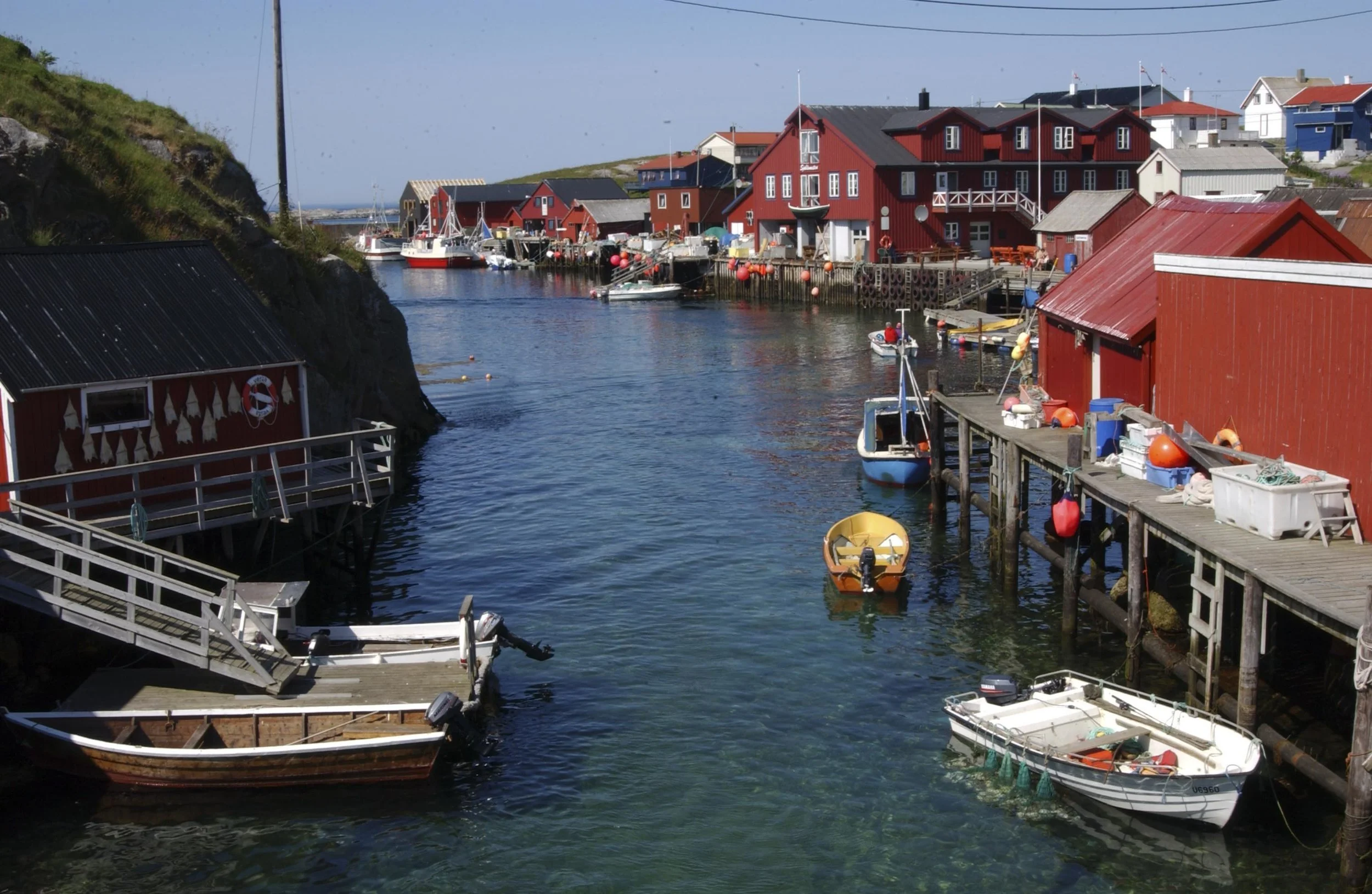 Et sund, med små røde naust på Mausund. Båter ligger ved kaien og dupper på en solfylt dag.