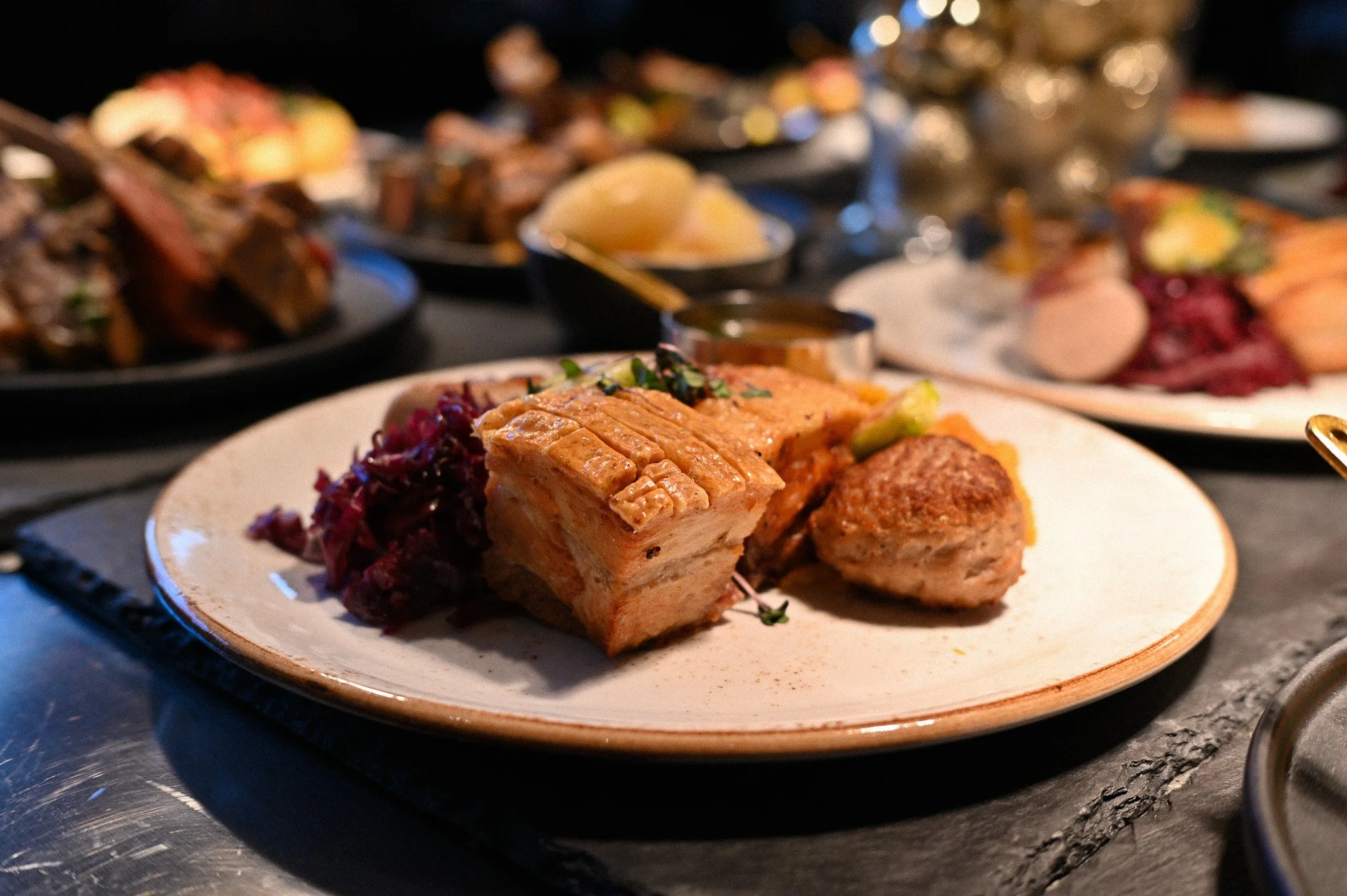 Nærbilde av en tallerken med ribbe, servert med rødkål, potetsalat og en liten skål med saus, på et serveringsfat. Bakgrunn med flere lignende retter.