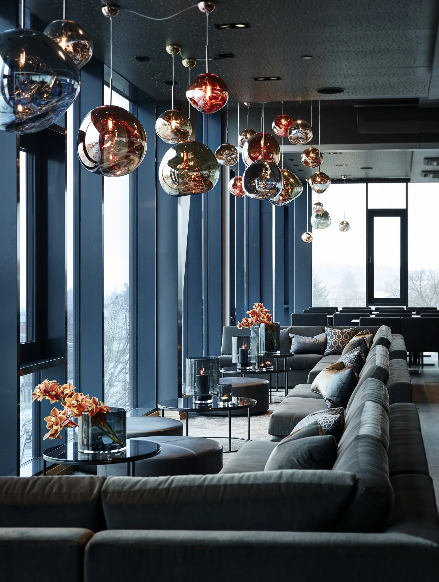 Moderne loungeområde med store vinduer, dekorative hengelamper i metall, blomsterdekorasjoner på svarte bord, komfortable sofaer og puter.
