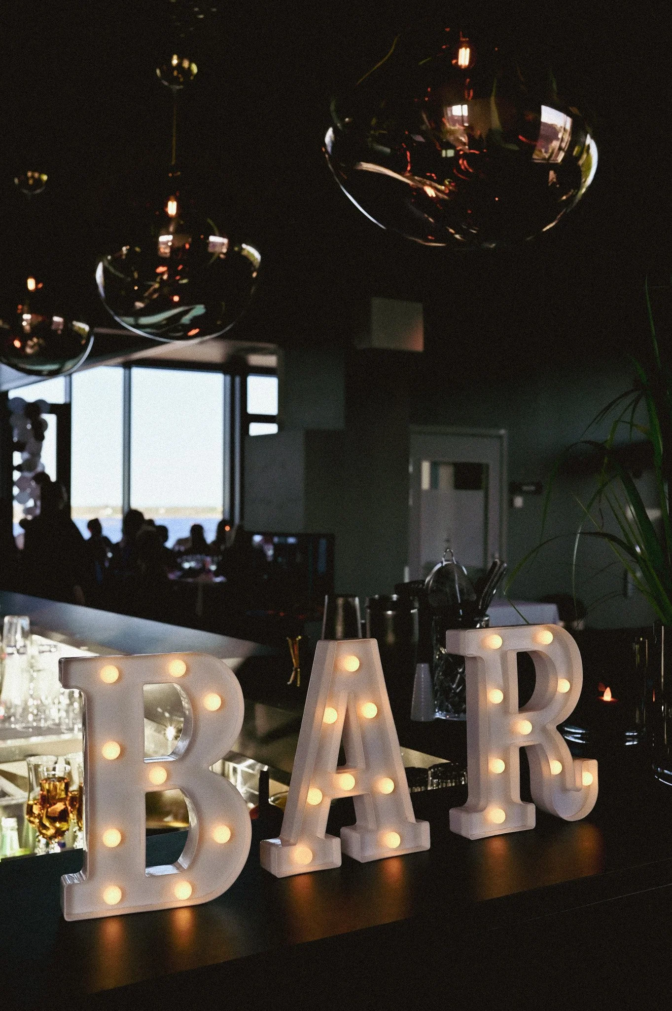 Innendørs bar med store opplyste bokstaver som skriver 'BAR' på en rekke, omgitt av glass og bartilbehør.