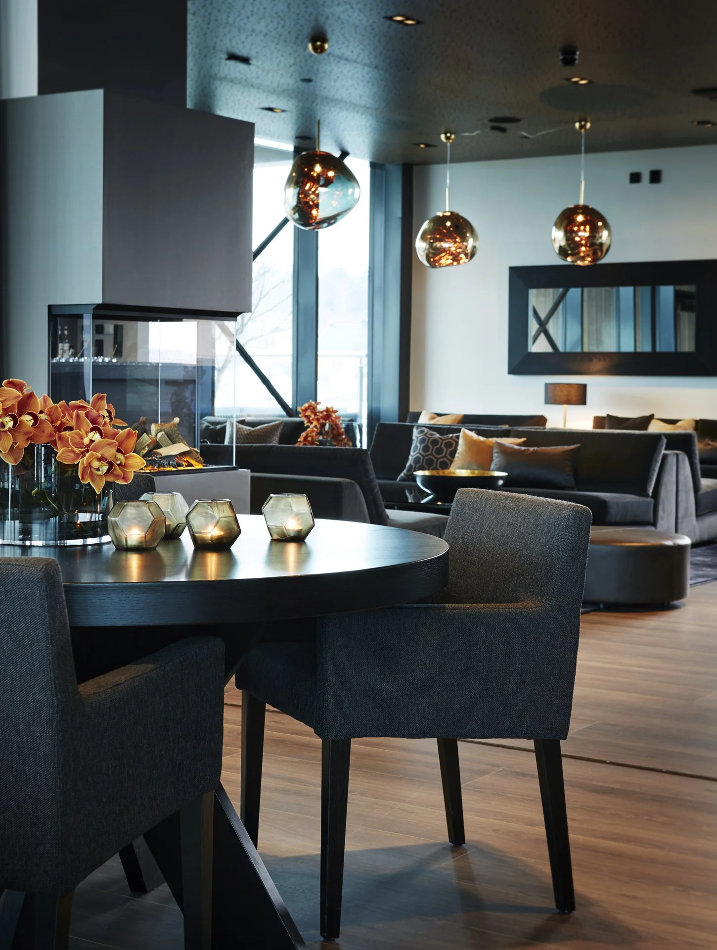 Moderne stue med svart spisebord, seks svarte stoler med stoffoverflate, blomster i vase, to lys i glasskåler, sofa med puter, store vinduer, tre kobberlamper i taket, speil på vegg, parkettgulv.