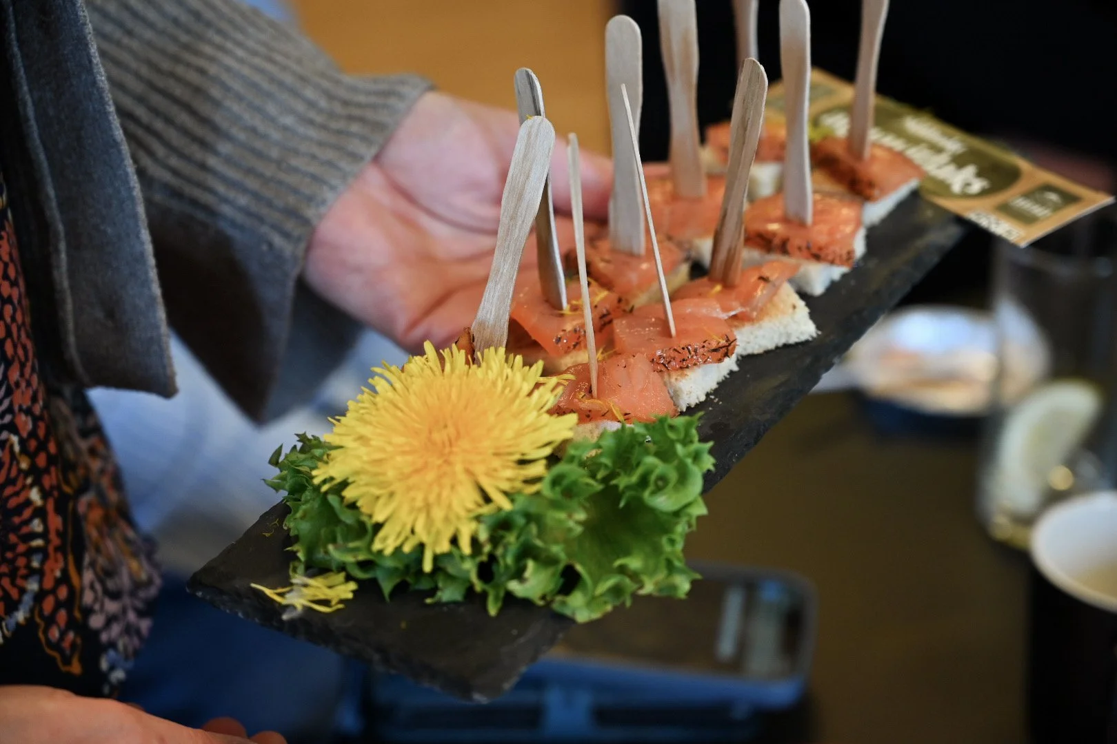 Et serveringsbrett av stein med smaksprøver fra Garnvik, røkt laks, loff og en gul løvetann som pynt