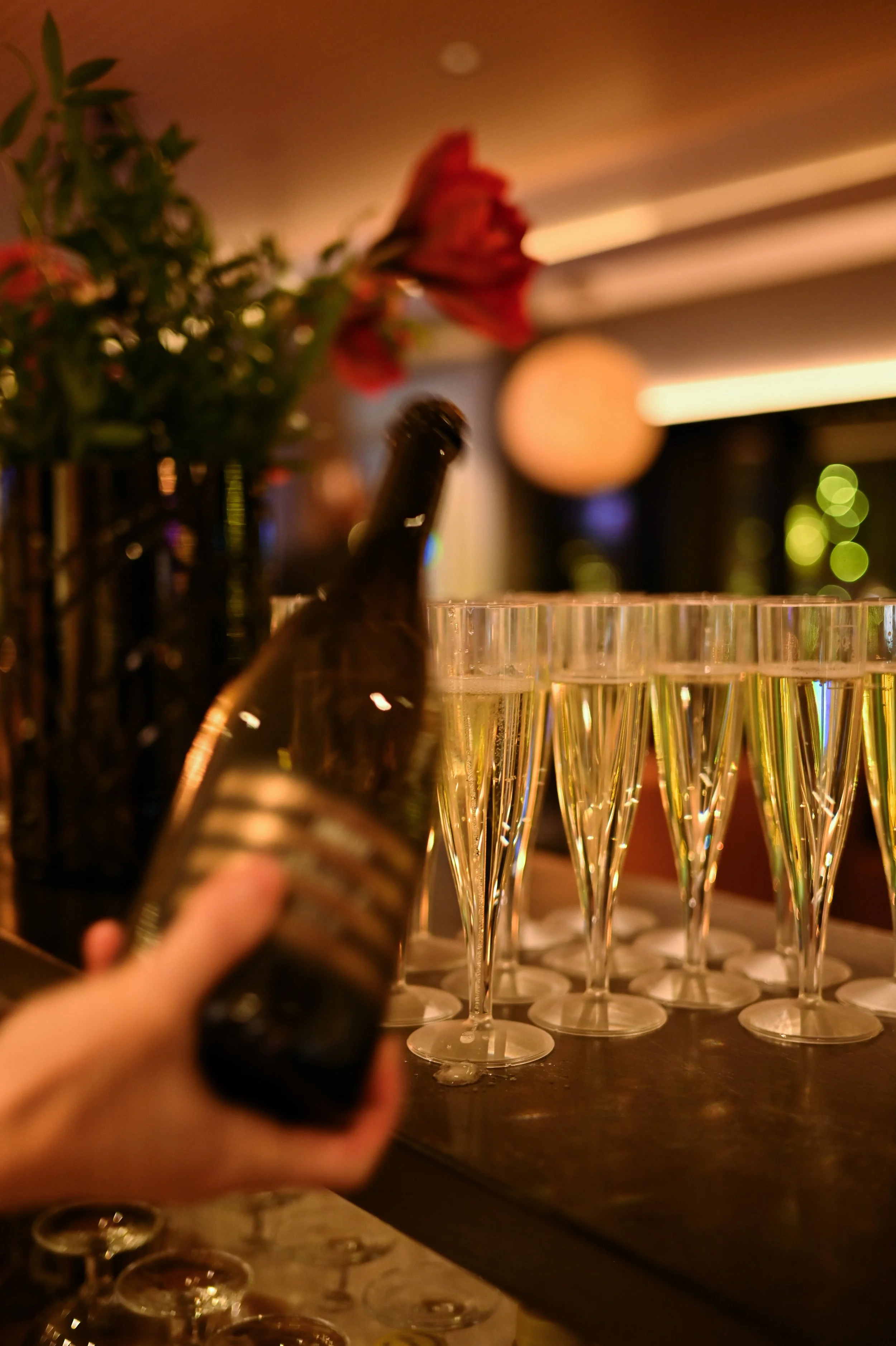 En person holder en flaske med musserende vin over champagneglass. I bakgrunnen er det flere tomme champagneglass og dekorasjoner, med varm belysning.