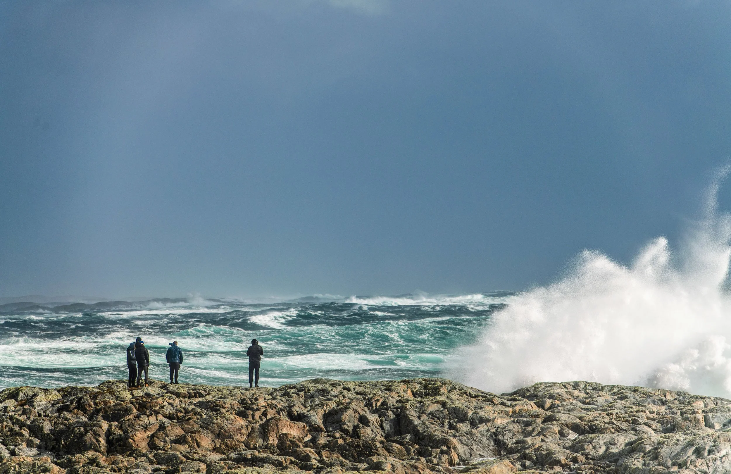 Fire personer som står i fjæra ved havet, en stor bølge raser til høyre i bildet. Beskriver været på Titran.