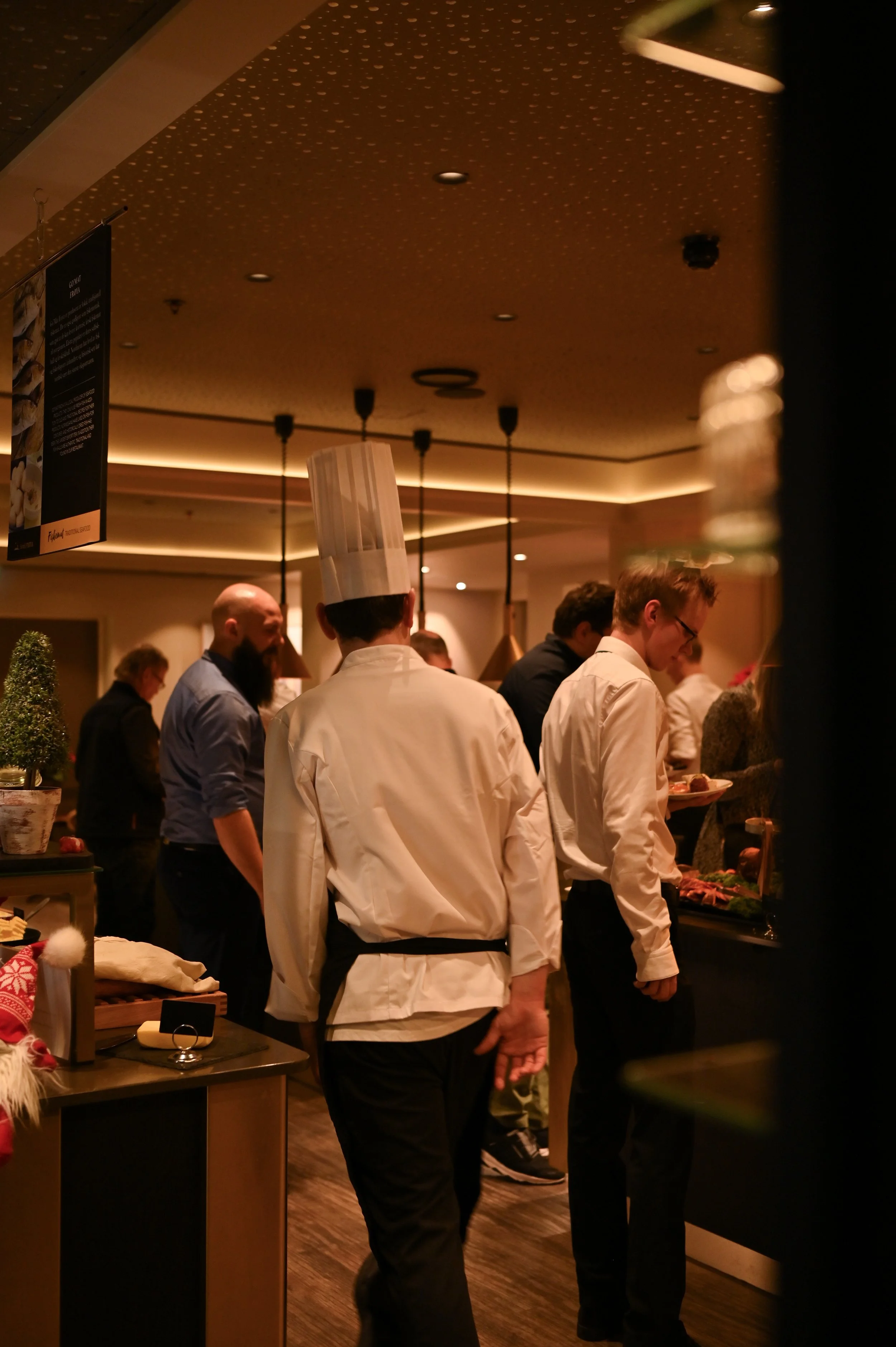 Bildet viser personer i en restaurant eller buffet, der noen av dem, inkludert en kokk i hvit uniform og kokkehatt, står sammen og velger mat fra en buffet. Det er dempet belysning med varme toner.