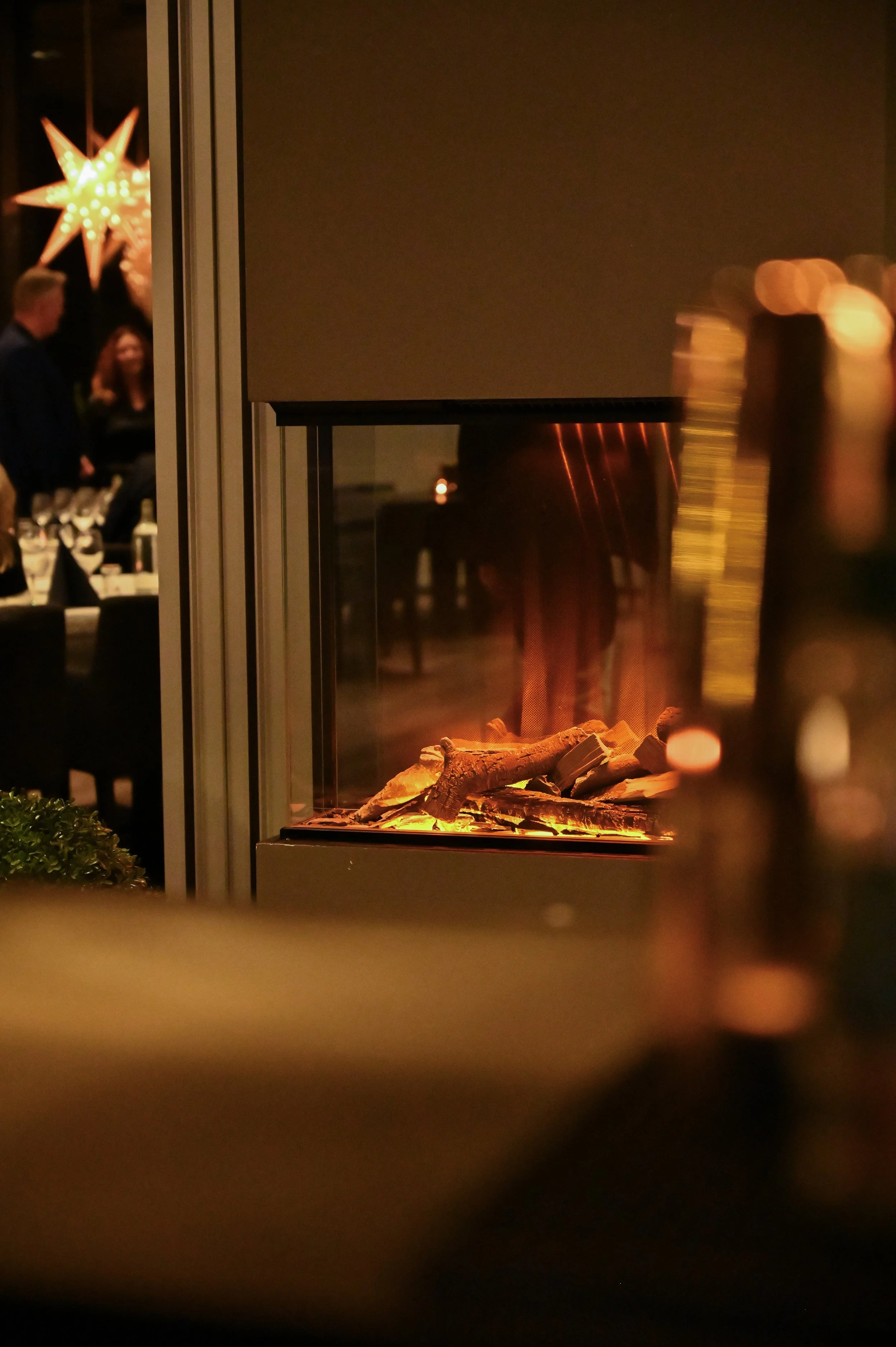 Innendørs peis med glødende kull og vedkubber, refleksjon av varme i glass. Bakgrunn med mennesker som snakker, og en stor dekorativ stjerne av lys.