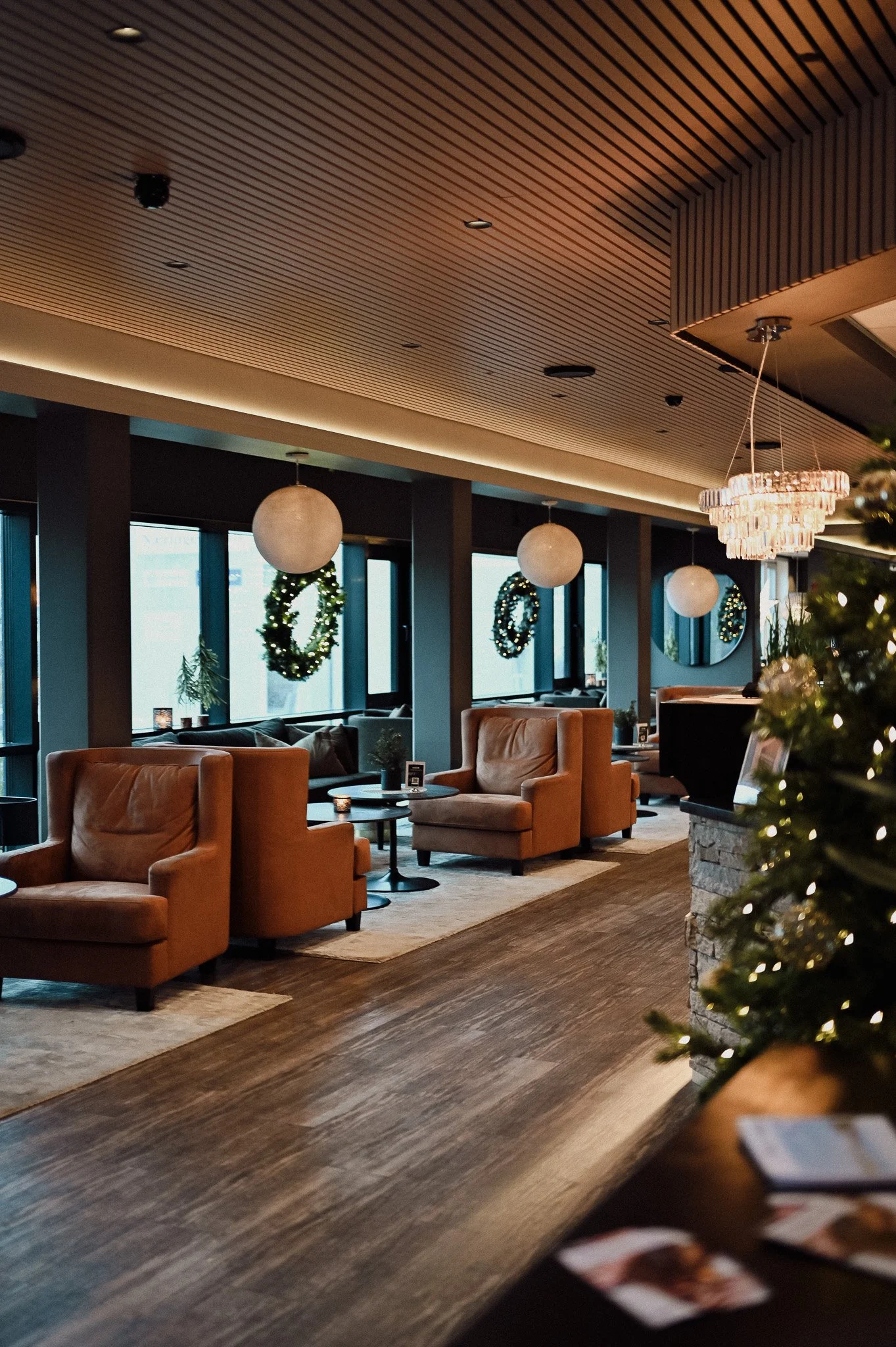 Innendørs område med lenestoler, små bord og juledekorasjoner, inkludert julekranser med lys, og ett speil på veggen. Det er store vinduer med utsikt utendørs.