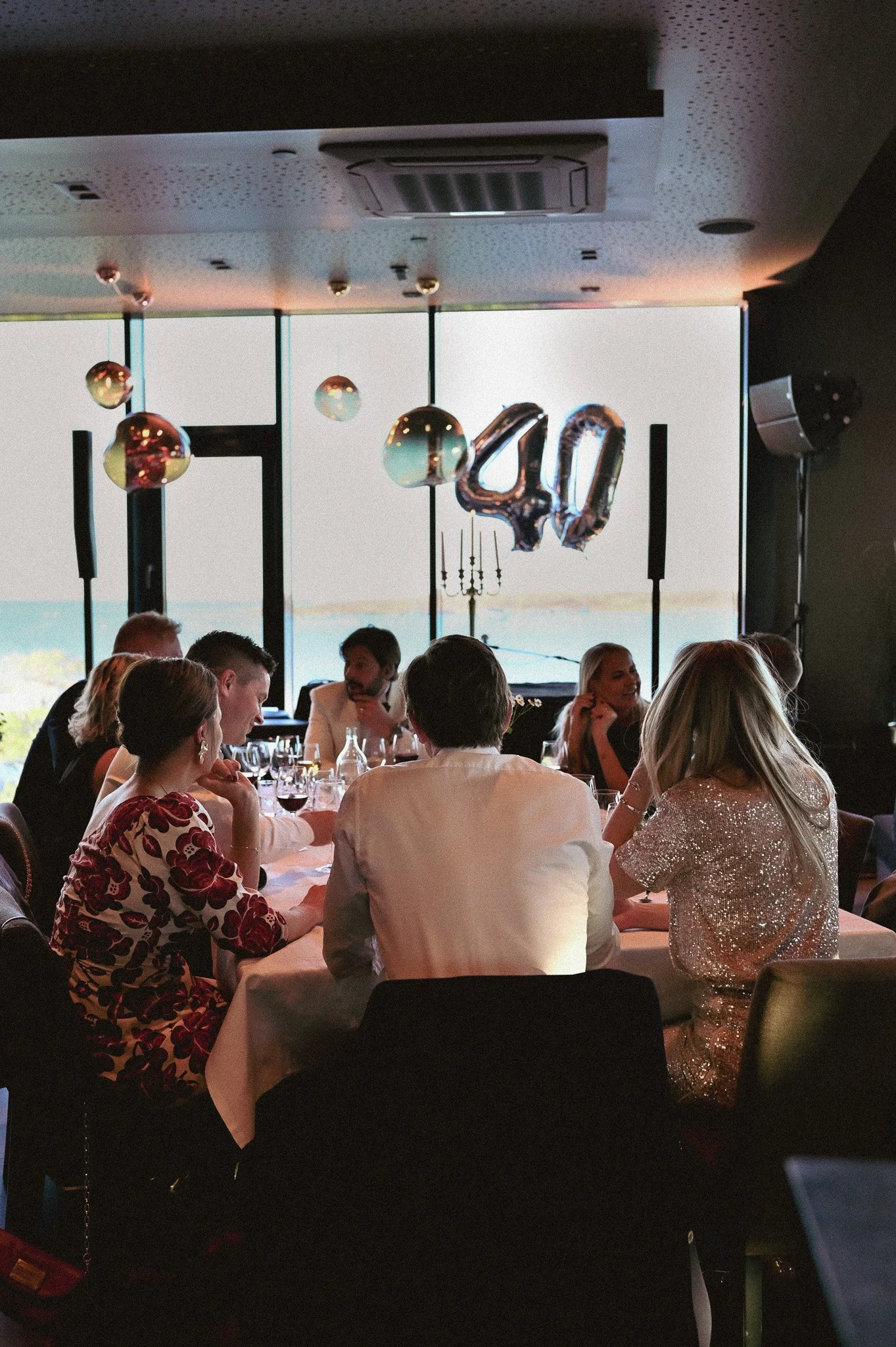Mennesker som sitter rundt et langbord og spiser, feirer 40-årsdag med sølvballonger og store folieballonger som viser tallet '40'.