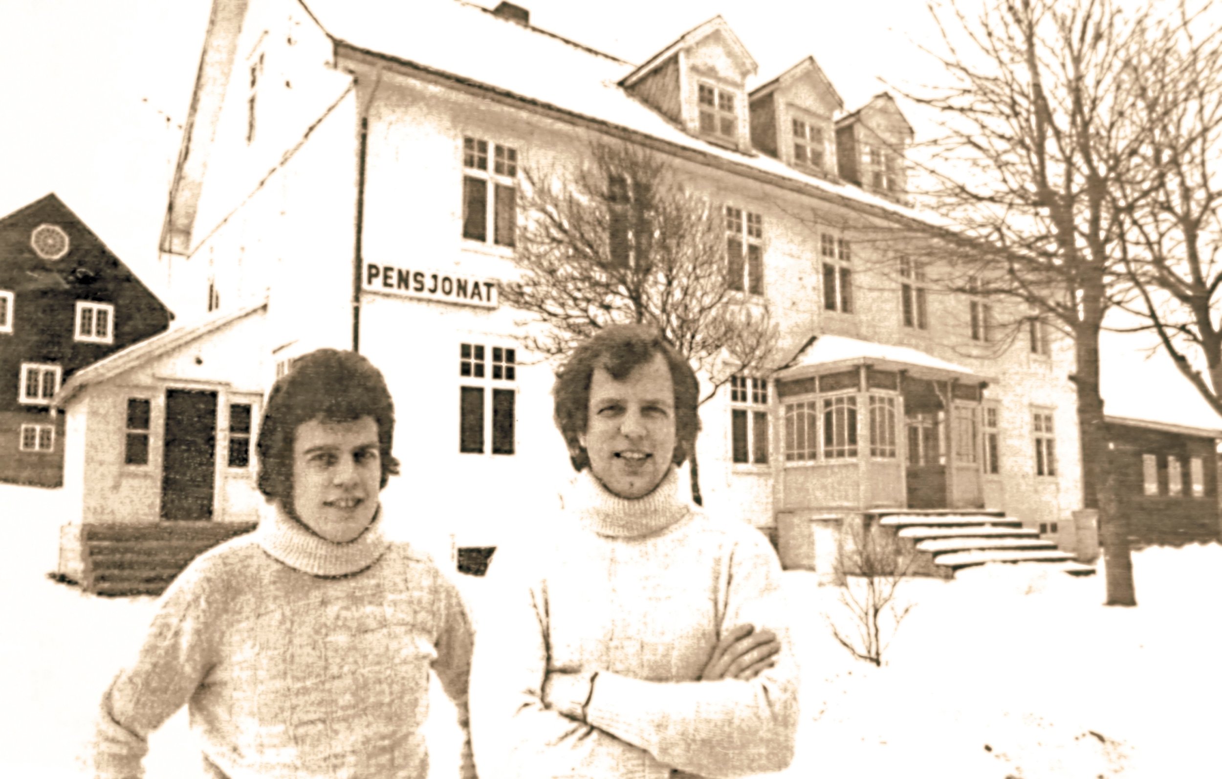 To unge menn står foran en stor, eldre bygning med skiltet "PENSJONAT". Det er snø på bakken, og trærne er uten blader, noe som antyder vinter.