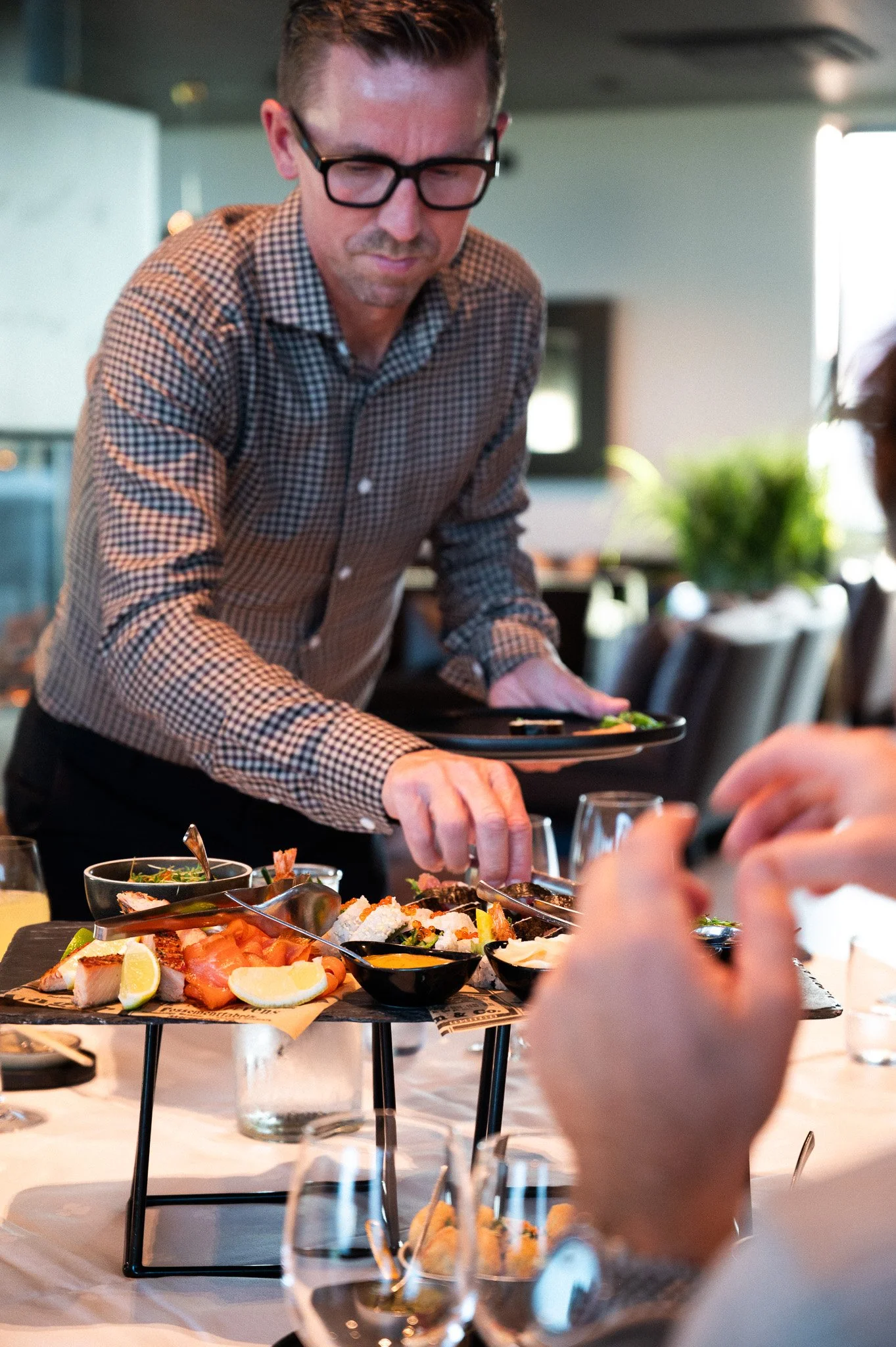 En mann i brunhvit rutete skjorte forskyner seg fra et fat med forskjellige matretter, blant annet røyket fisk, sitron og sushi. Fatet er plassert på et flott dekket bord med hvit duk.