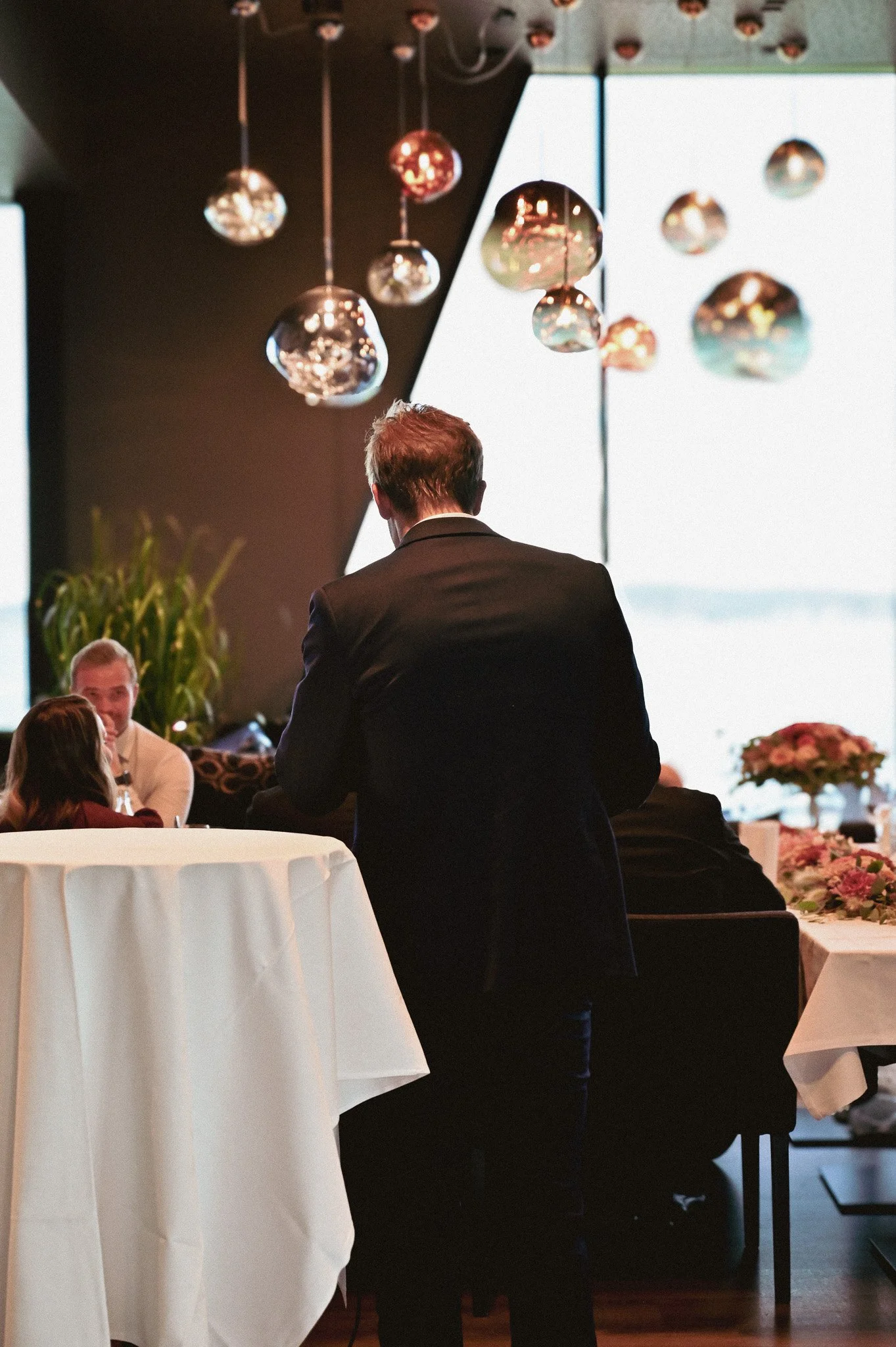 En mann i svart dress står ved et rundt bord under et elegant selskap, med flere andre personer i bakgrunnen, og moderne lyselementer henger fra taket.