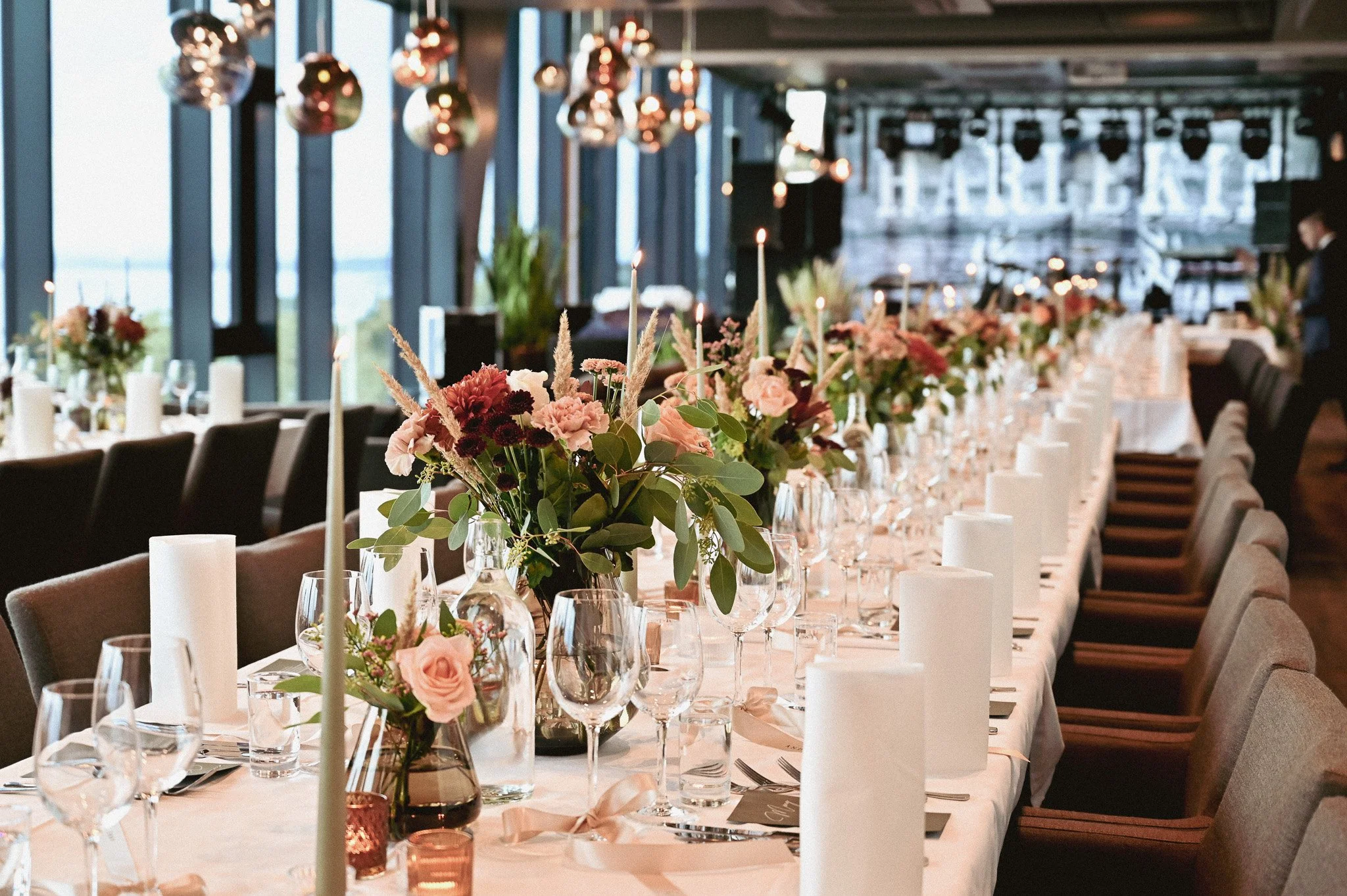 Langbord dekket for et formelt måltid med blomsterarrangementer, lys, glass og bestikk i et moderne rom med store vinduer.