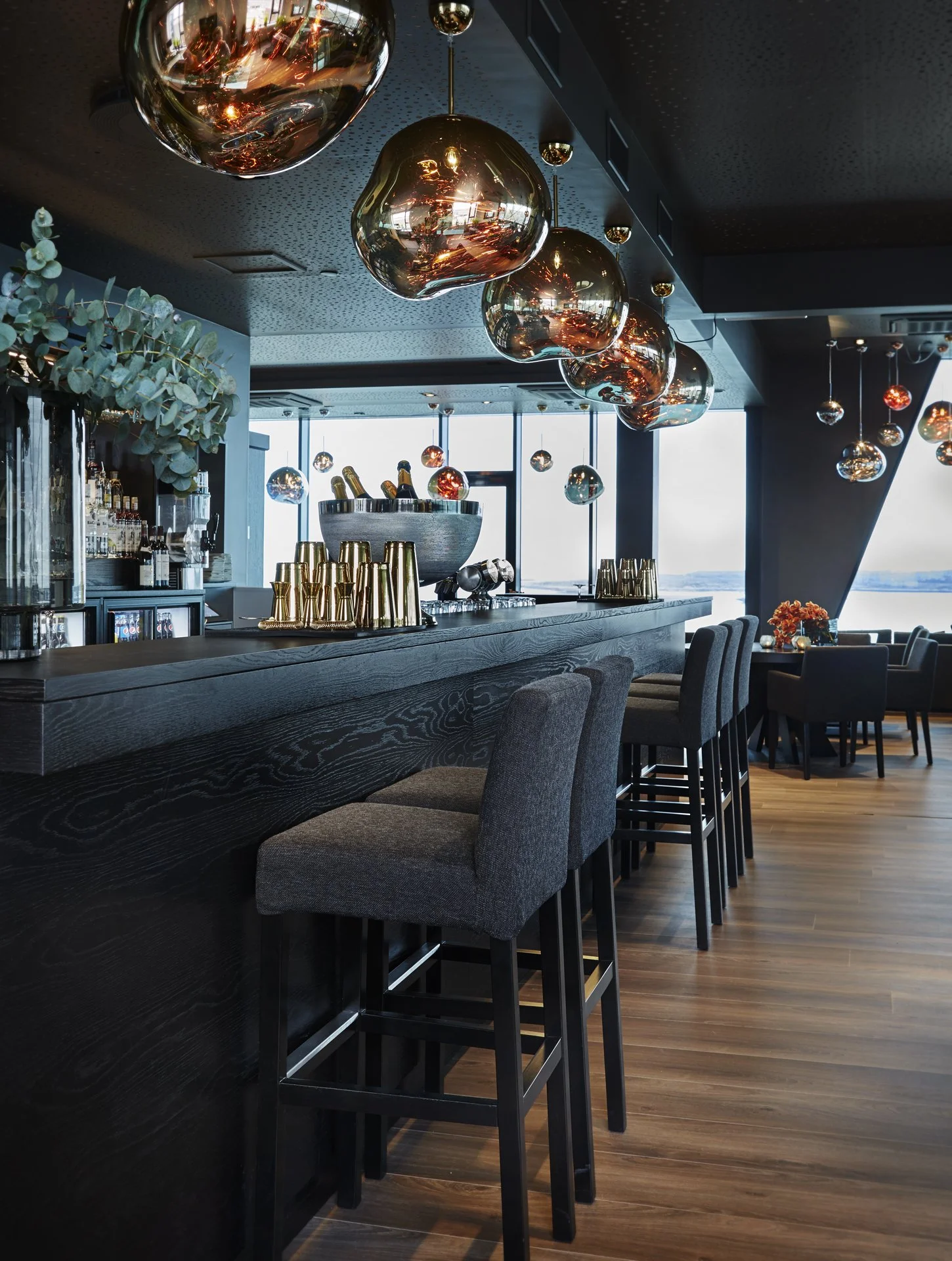 Interiør av en bar eller restaurant med høye stoler, dekorative hengende lamper og utsikt gjennom store vinduer.