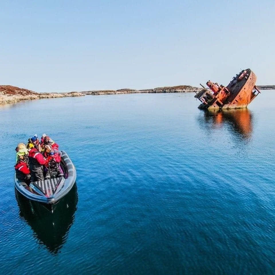 En gruppe turister på en båt ser på et sunket, rustent skip som står på siden i vannet.