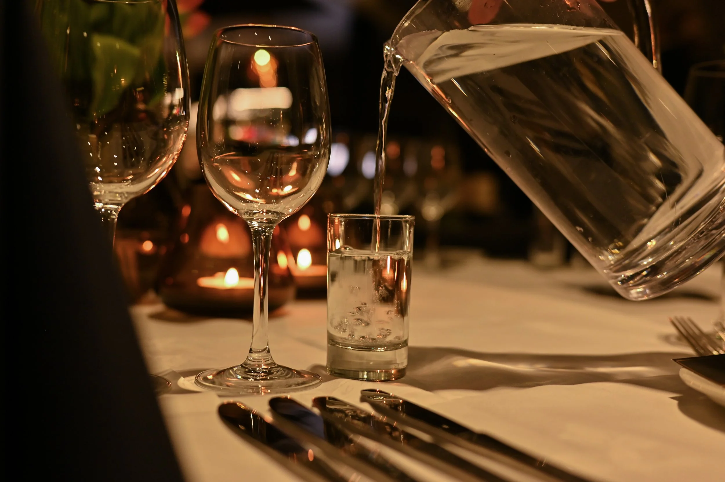 Nærbilde av et vinglass, et lite glass med vann, med en bakgrunn av lys og glass. Vann blir hellet fra en stor glasskaraffel.