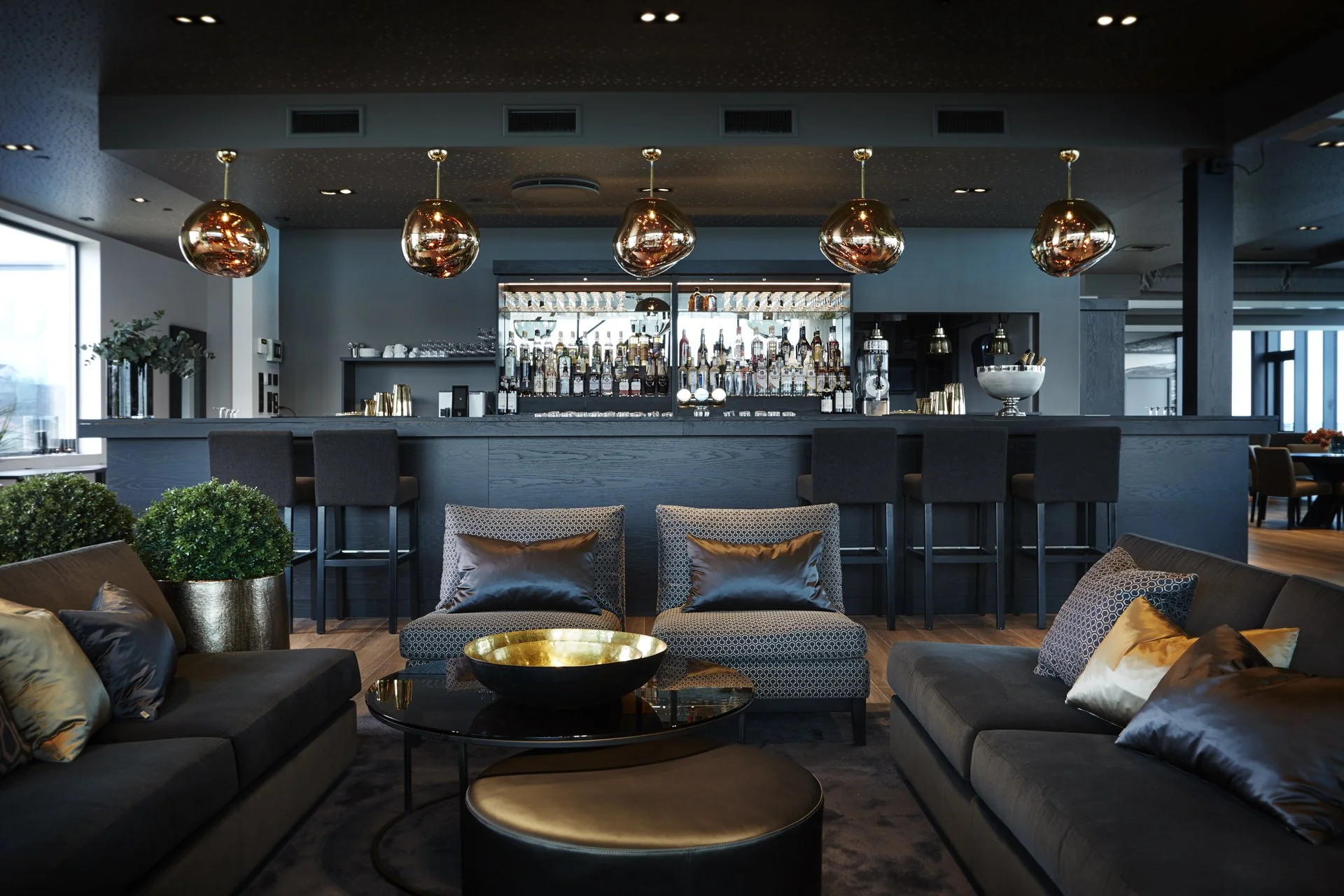 Innendørs hotellbar med sitteplasser, en bar med bakgrunn av alkoholflasker, og moderne dekorasjoner i gull og svart.