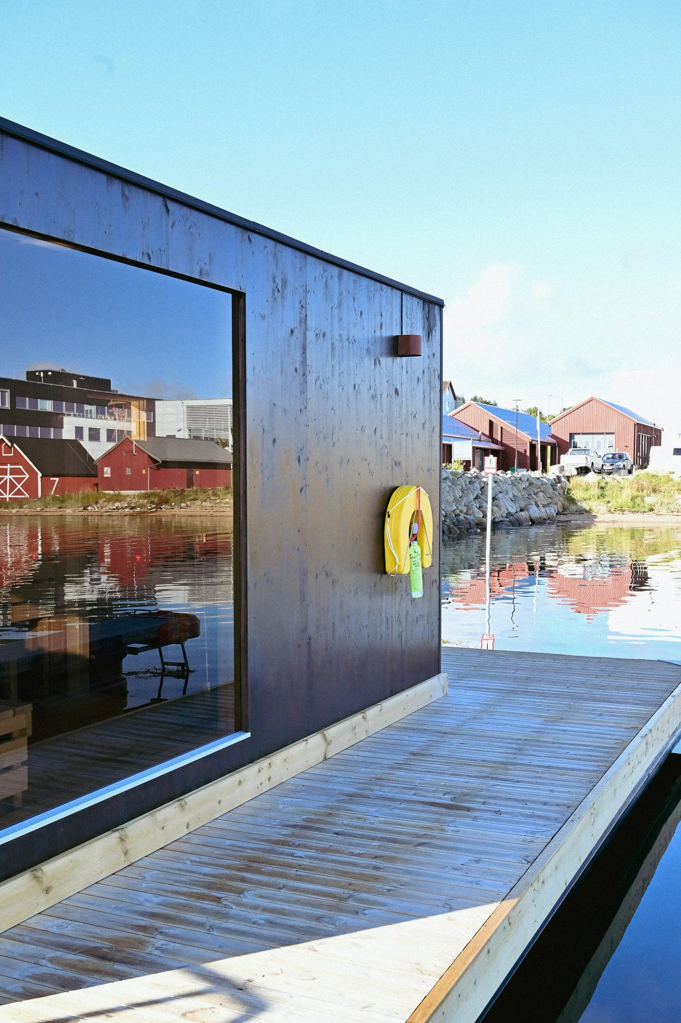 Bilde av en trebelagt brygge med Badstue ved vann med moderne bygninger og båter i bakgrunnen.