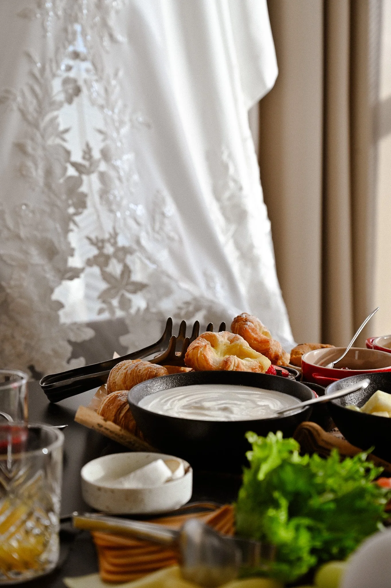 Et bord med frokostmat som croissanter, yoghurt, grønnsaker og andre småretter, med en hvit brudekjole og gardiner i bakgrunnen.