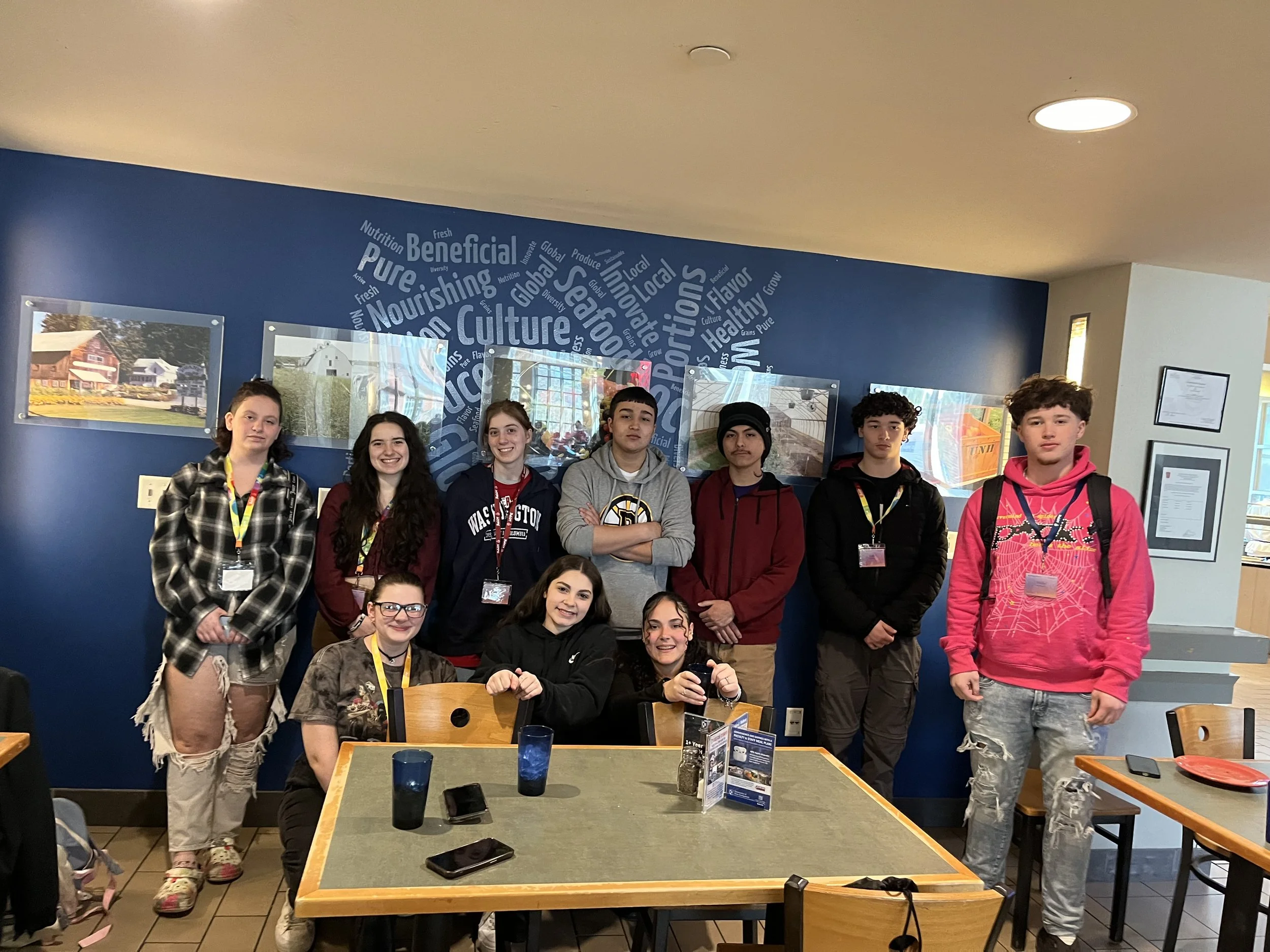 Group of ten teenagers, six standing and four sitting, posing inside a restaurant with blue wall decorated with words and pictures. Some are smiling, some with neutral expressions. Dining table in foreground with phones and glasses.