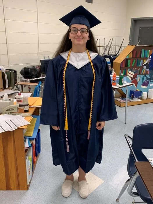 A young woman in a navy blue graduation cap and gown, wearing a white collar and a gold honorary rope, standing in a classroom with bookshelves, desks, and various supplies.