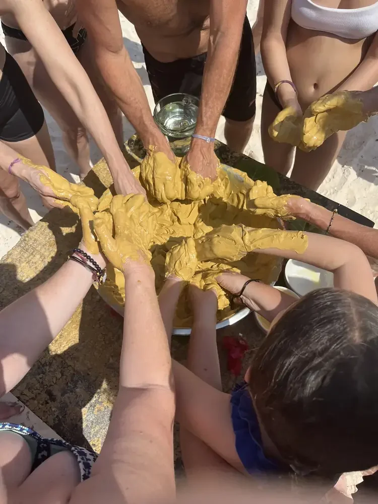 Hands reaching into a shared bowl of clay on the beach.