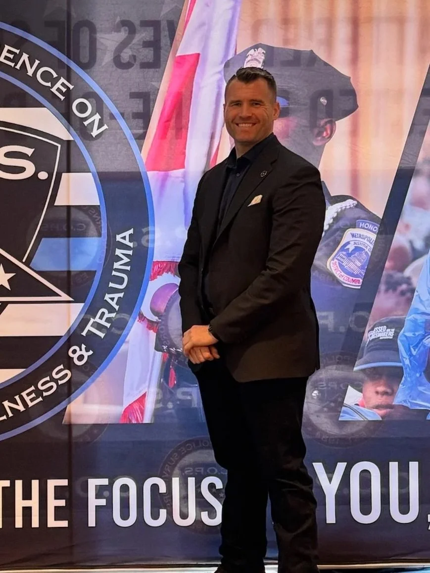 A man in a black suit smiling and standing in front of a promotional backdrop with police-related imagery and logos.