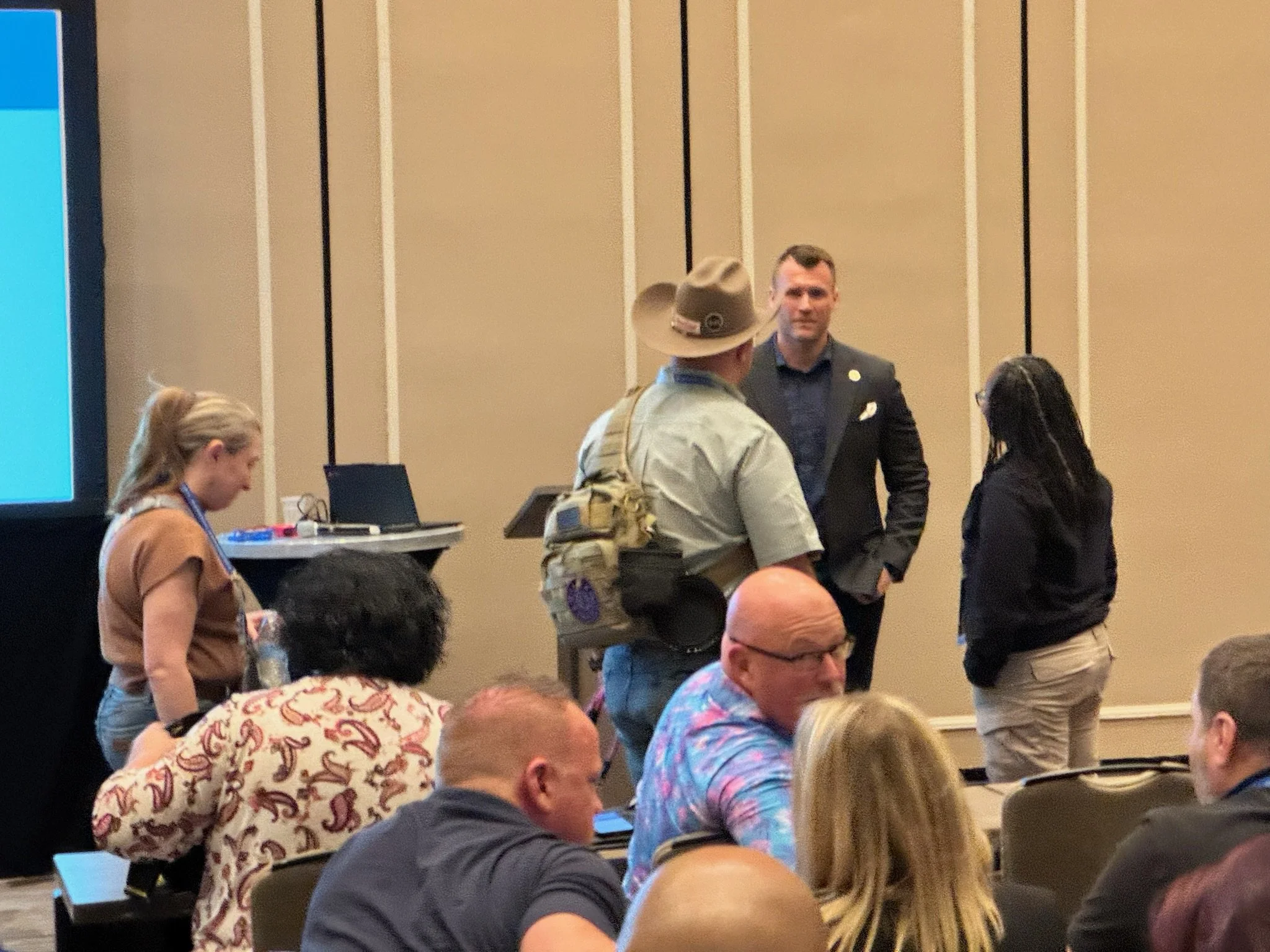 Group of people standing and sitting in a conference room, engaged in conversation, with a presentation screen on the left side.